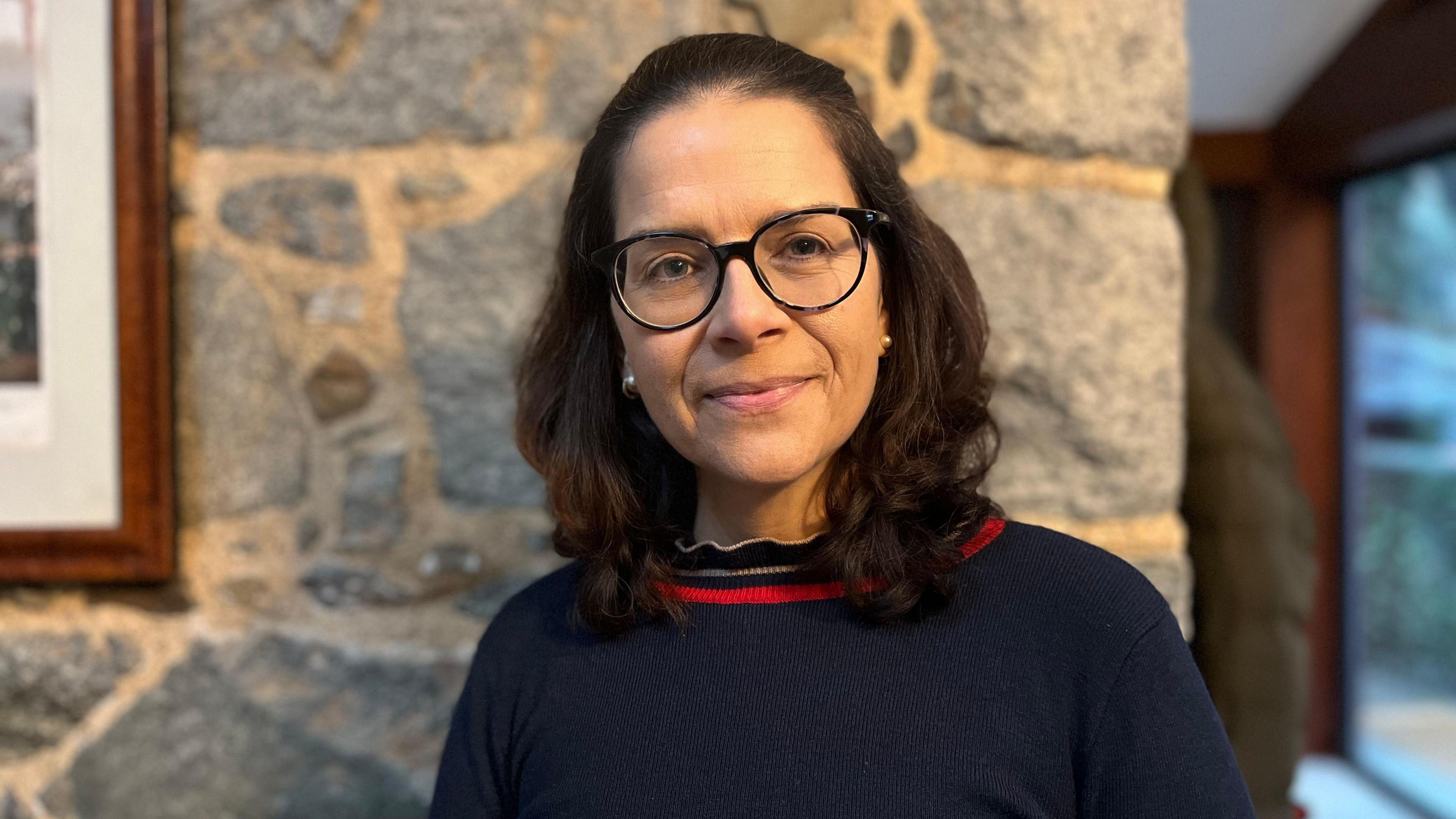 A middle-aged woman in a blue jumper. She has shoulder length hair and glasses and stands by an interior granite wall with the corner of a picture frame visible.