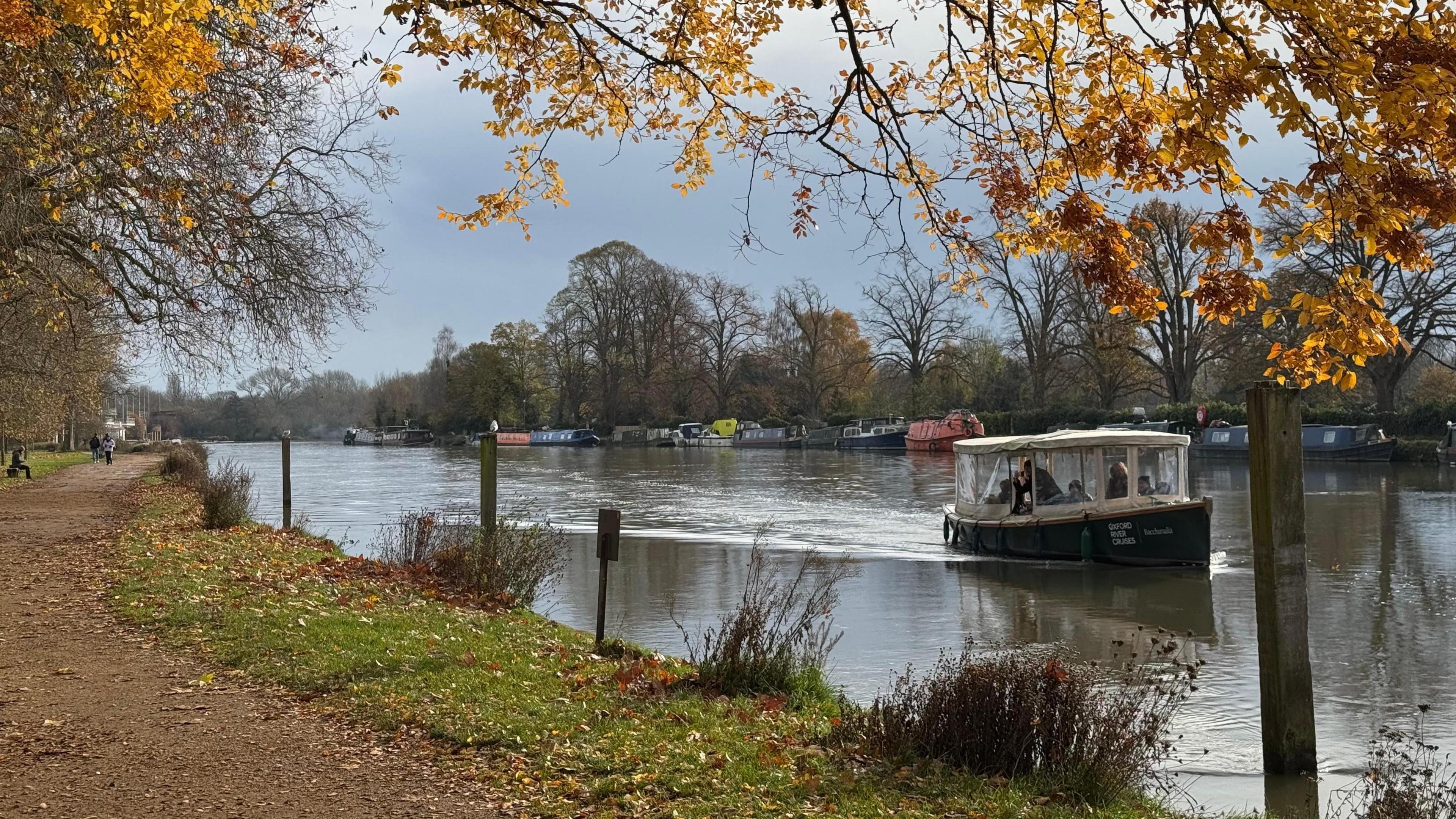 A river scene with a motorboat sailing on the water. Trees can be seen lining the river with more boats moored on the far side. The sky is grey and looking threatening.