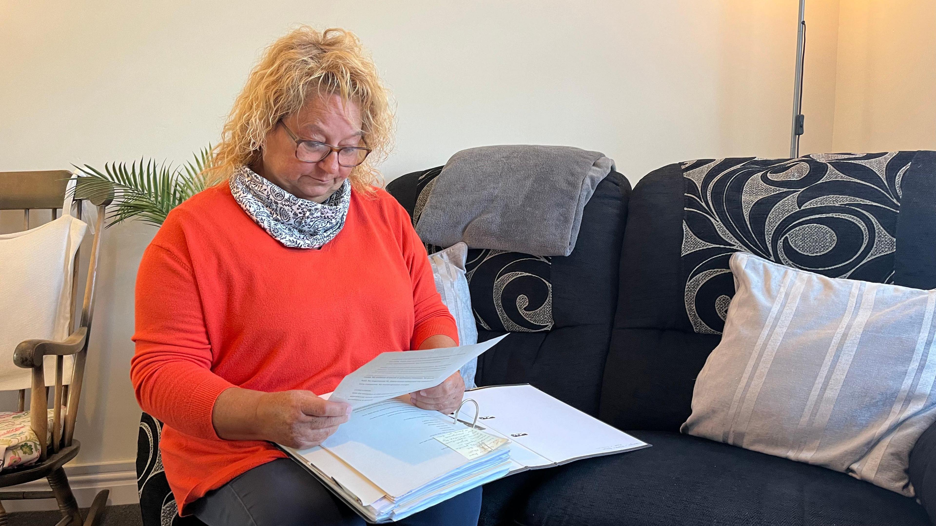 A woman in a white, paisley-patterned scarf and orange jumper sat on a black sofa while looking at a piece of paper with an open folder filled with paper on her lap