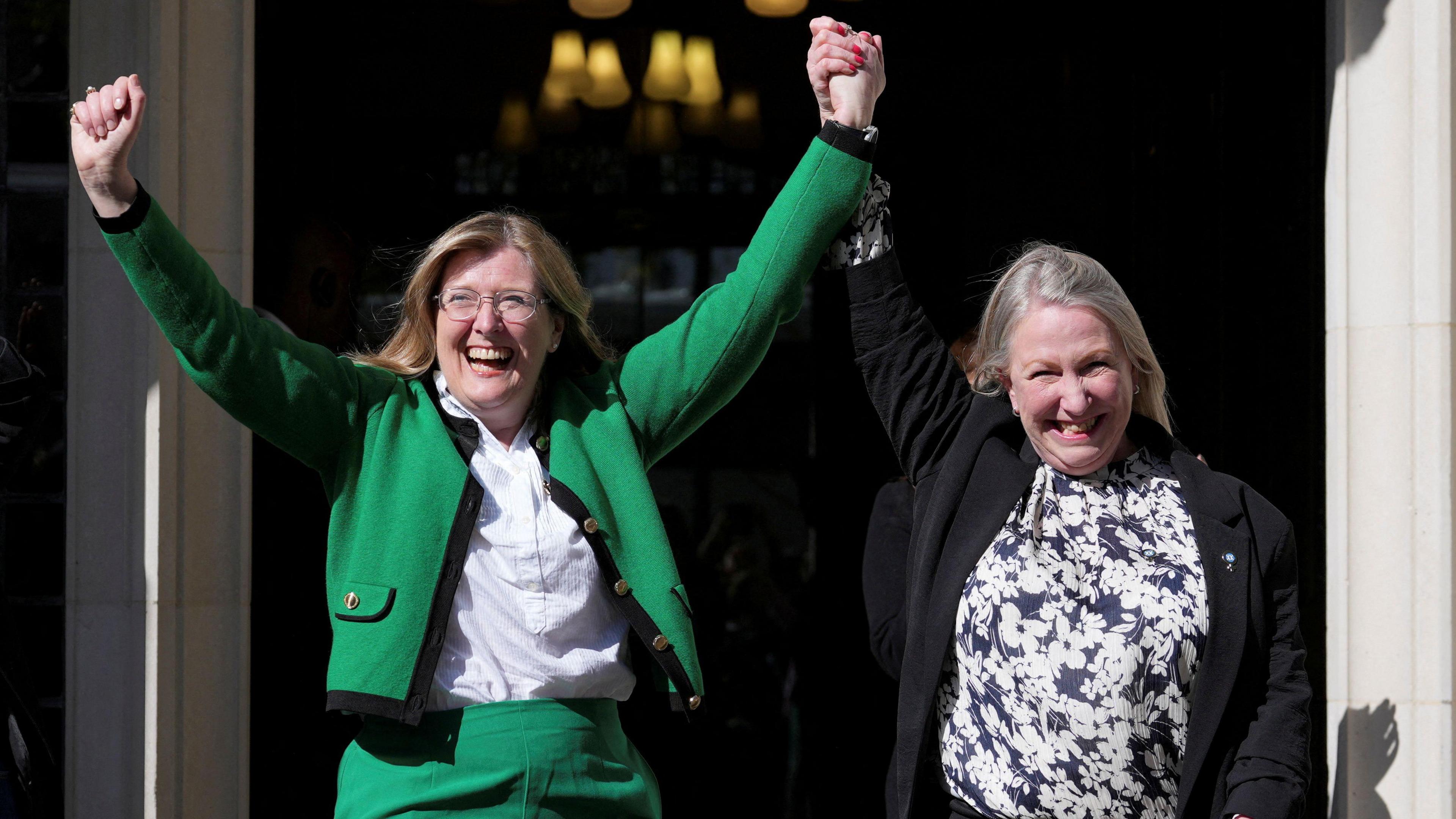 Two women celebrating outside the Supreme Court. The woman on the left is wearing a green suit with the jacket open over a white shirt. She has her hands in the air. Her left arm his being raised by the woman on the right, who has her right arm in the air. She is wearing a black jacket, which is open, over a blue and white floral patterned top.