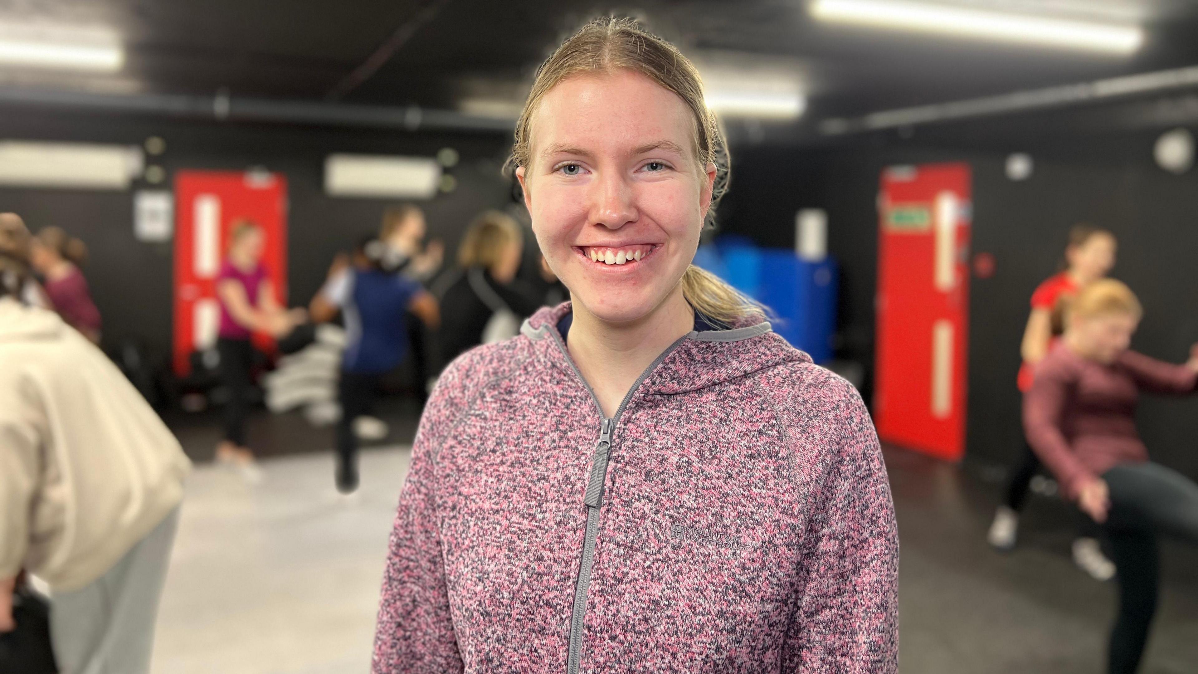 Lucy smiles at the camera while girls train behind her in the room. She is wearing a pink and grey hoodie and she has blue eyes and long blonde hair which is tied into a pony tail.