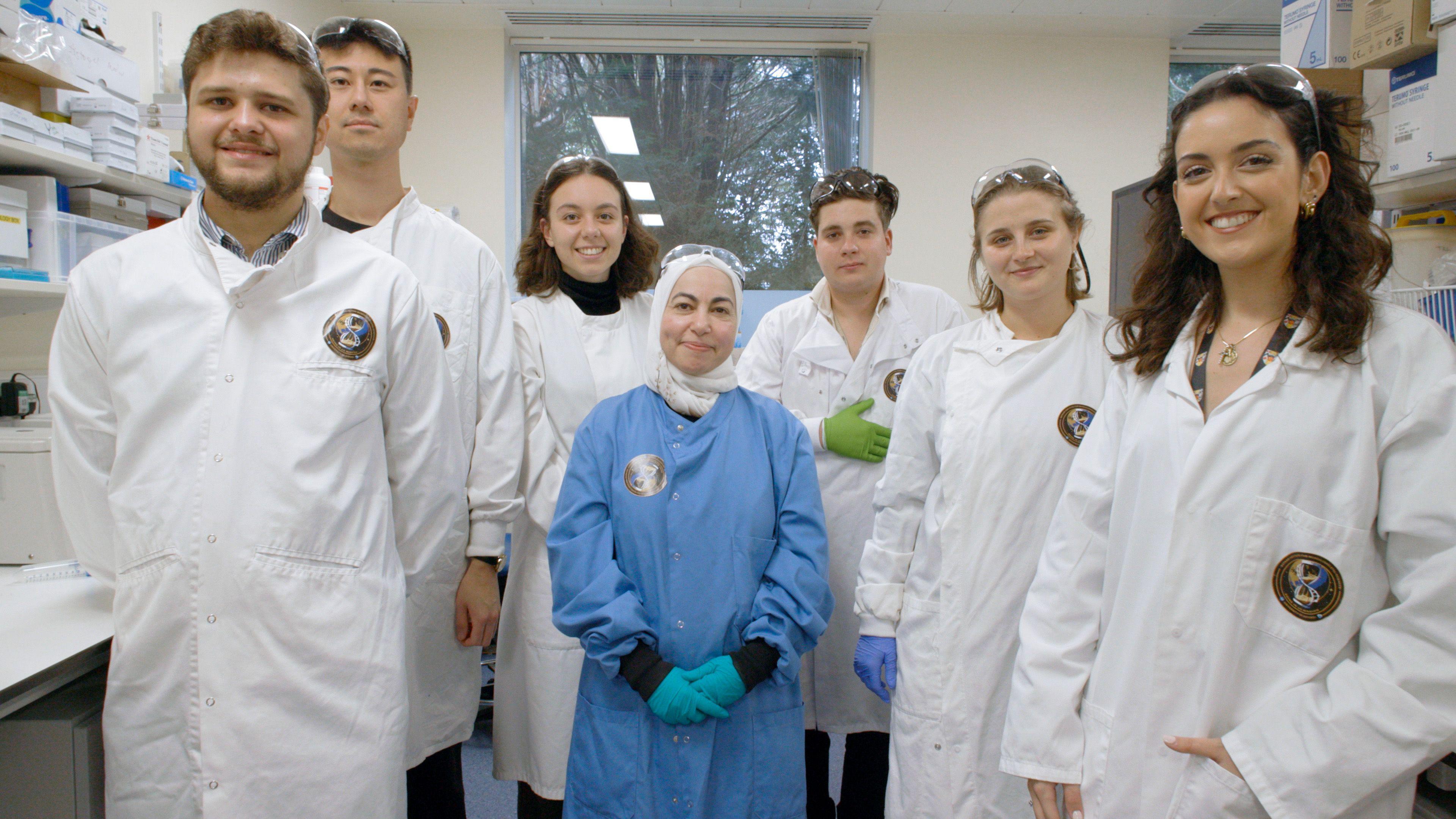 Dr Ghada Alsaleh and six researchers are smiling for the camera. She is in the middle wearing a blue coat. The researchers are all wearing white coats.