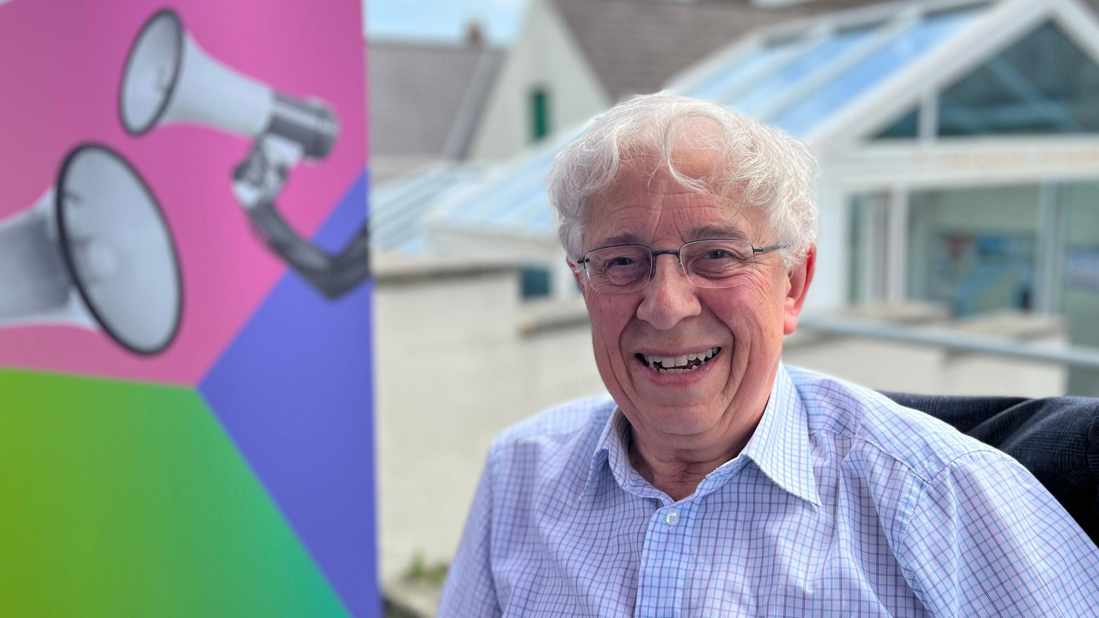 A man with white hair and glasses, wearing a blue and white checked shirt. On the left of the picture is the Your Voice, Your Vote logo. 