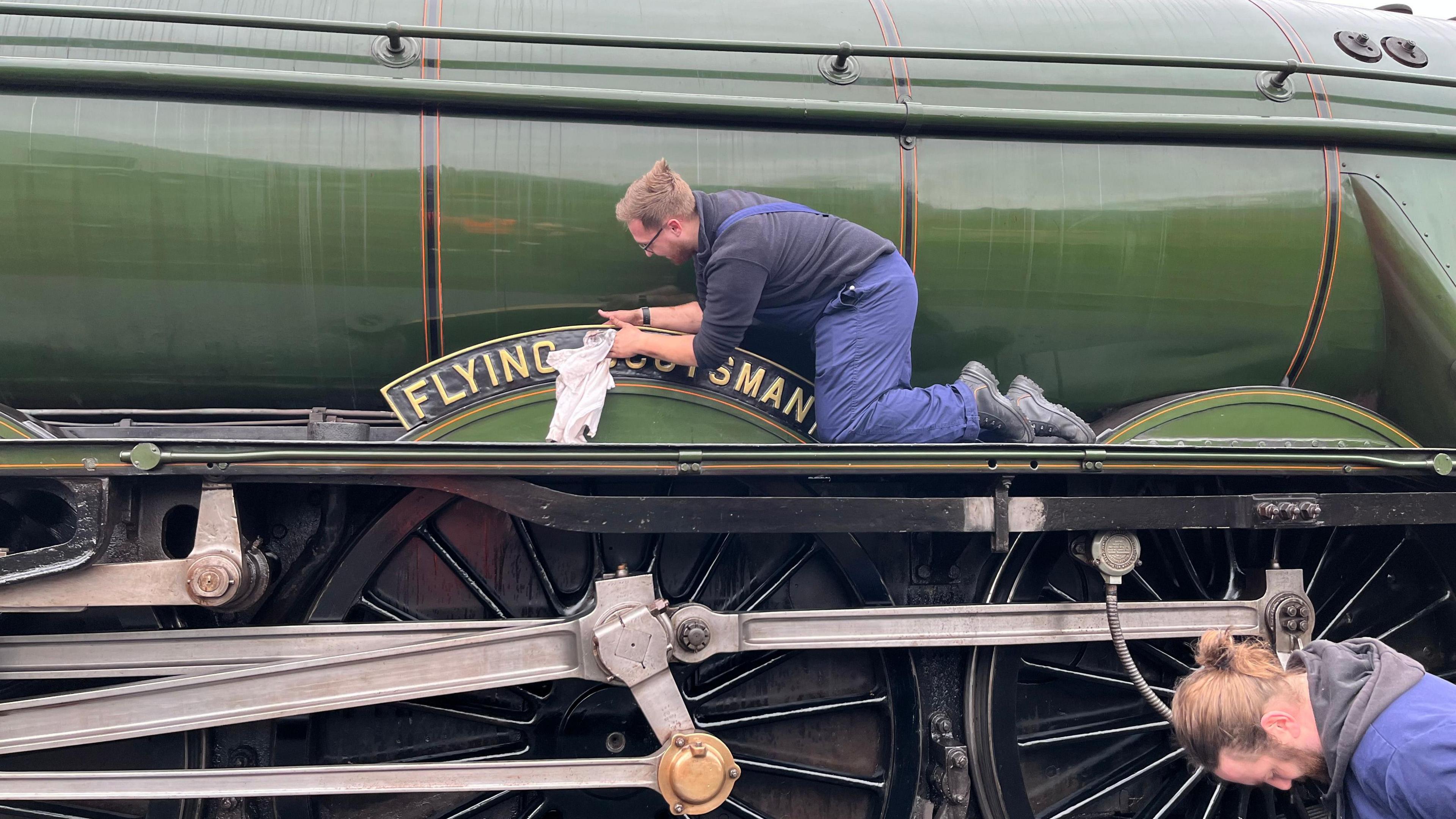 Green locomotive with a gold and black name plate - with two men in blue overalls- one on top of the train and one near its wheels cleaning it