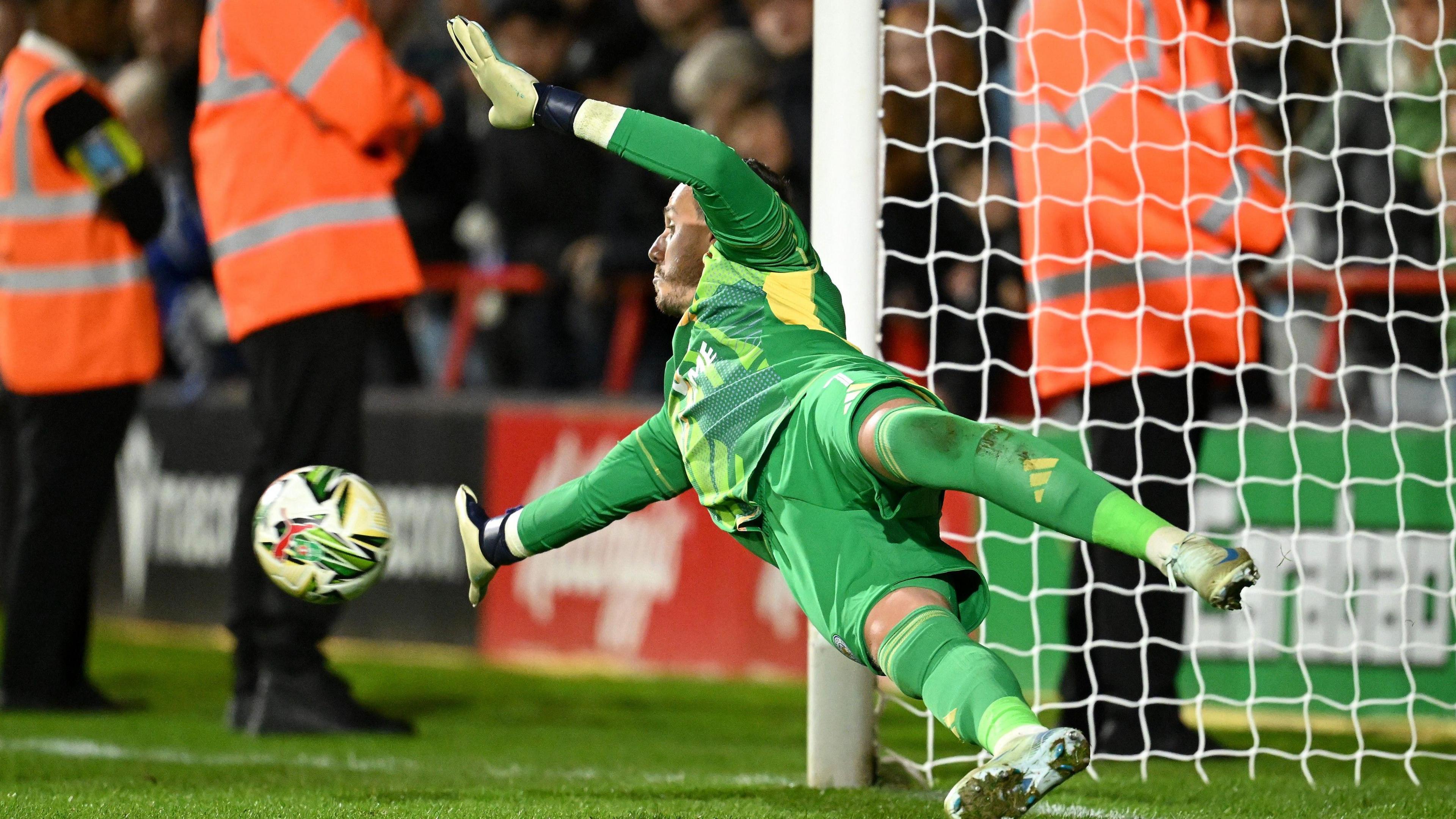 Danny Ward, wearing Leicester's green goalkeeping kit, dives to his right to save a low penalty with his right hand from Walsall player Taylor Allen (not pictured).