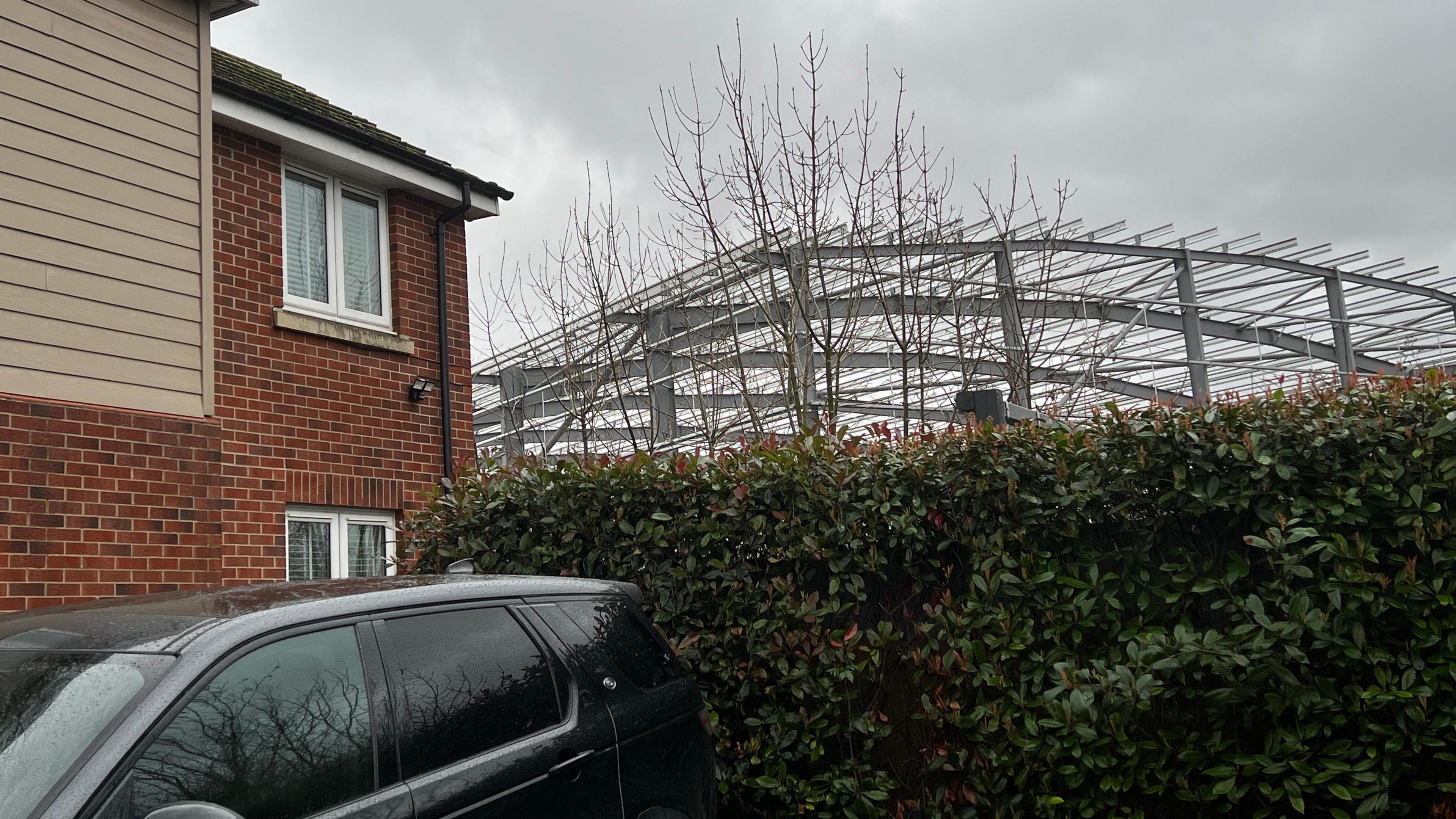 The corrugated metal structure of the warehouse can be seen looming over the top of a resident of Hooke Close's hedge next to their property. The resident's black car is also in shot by the red brick house. 