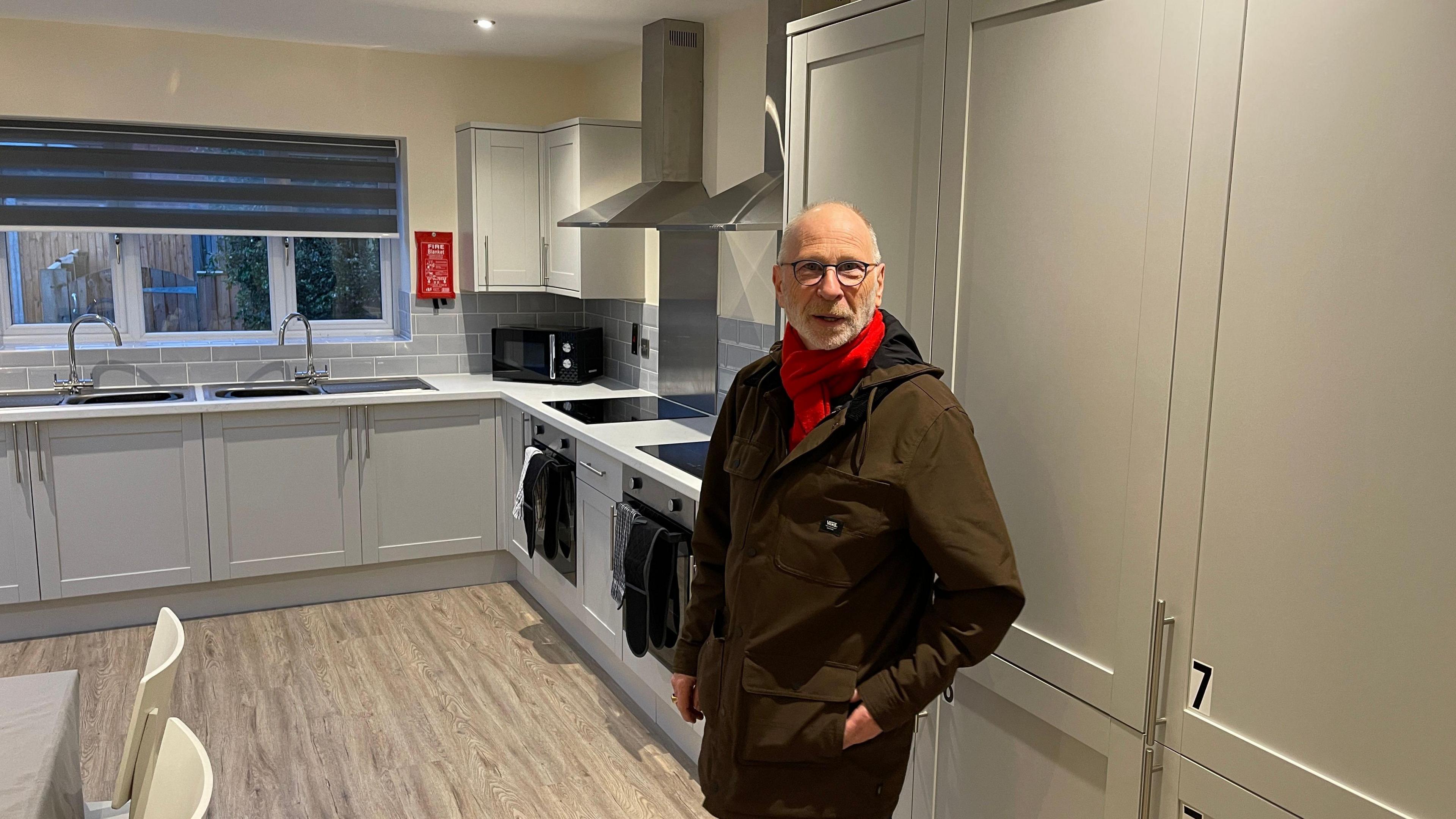 Richard O'Driscoll is standing in a kitchen of a new house, he is wearing a brown jacket and dark trousers. He has a red scarf wrapped around his neck. He is also waring glasses. 