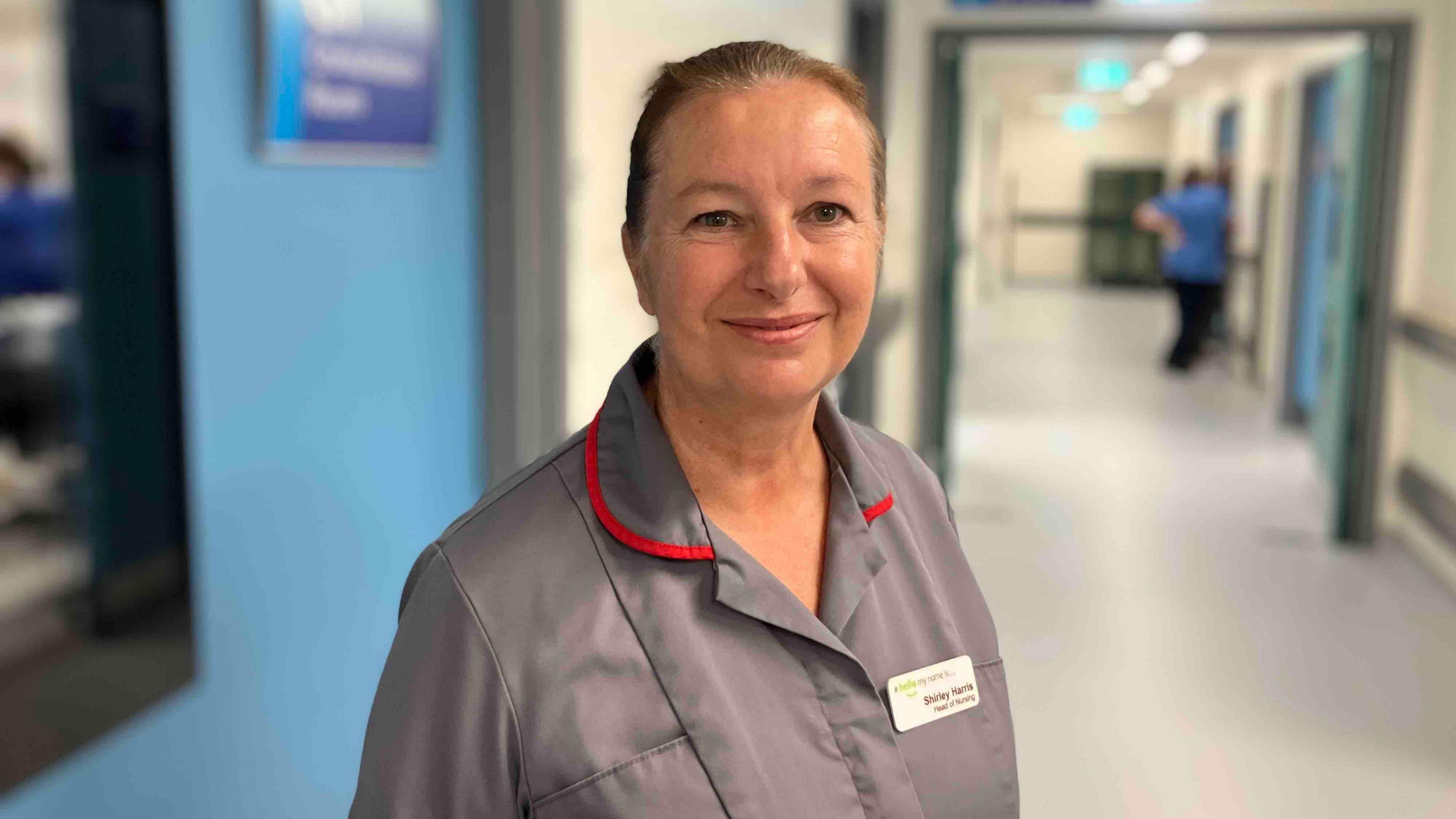 A nurse, looking at the camera and smiling. She has her hair tired back. She is wearing a grey nursing uniform and name badge. In the background is a corridor.