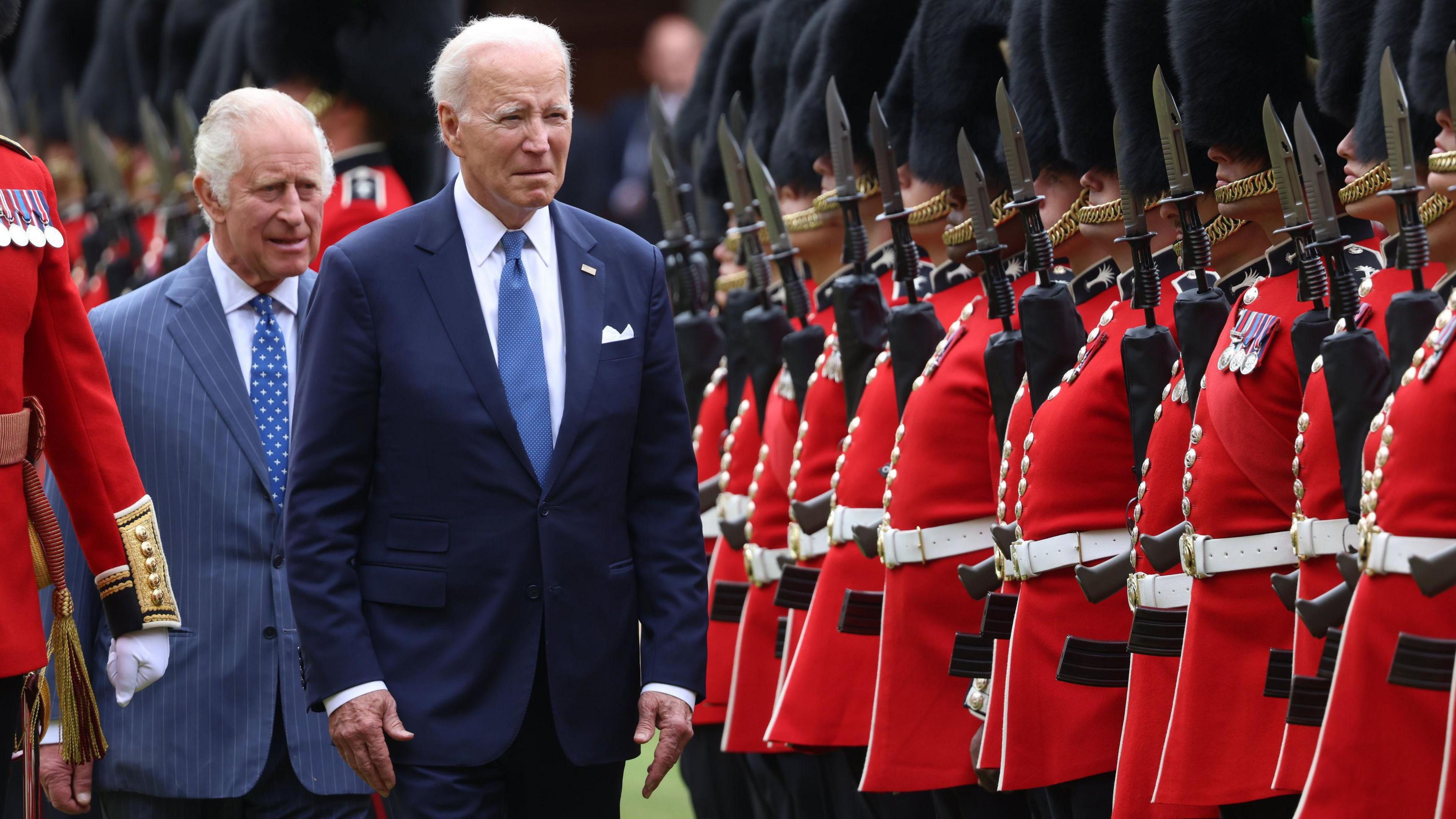 King Charles III and US President Joe Biden inspect the Guard of Honour from the Prince of Wales's Company of the Welsh Guards in the quadrangle at Windsor Castle, Berkshire, during President Biden's visit to the UK. Biden is walking in front of the king and is wearing a dark navy suit, white shirt and blue tie, King Charles, a lighter blue pinstripe suit, white shirt and blue tie. The Welsh Guards are lined up on the righthand side, wearing red uniforms and bearskin fur hats, and holding bayonets.