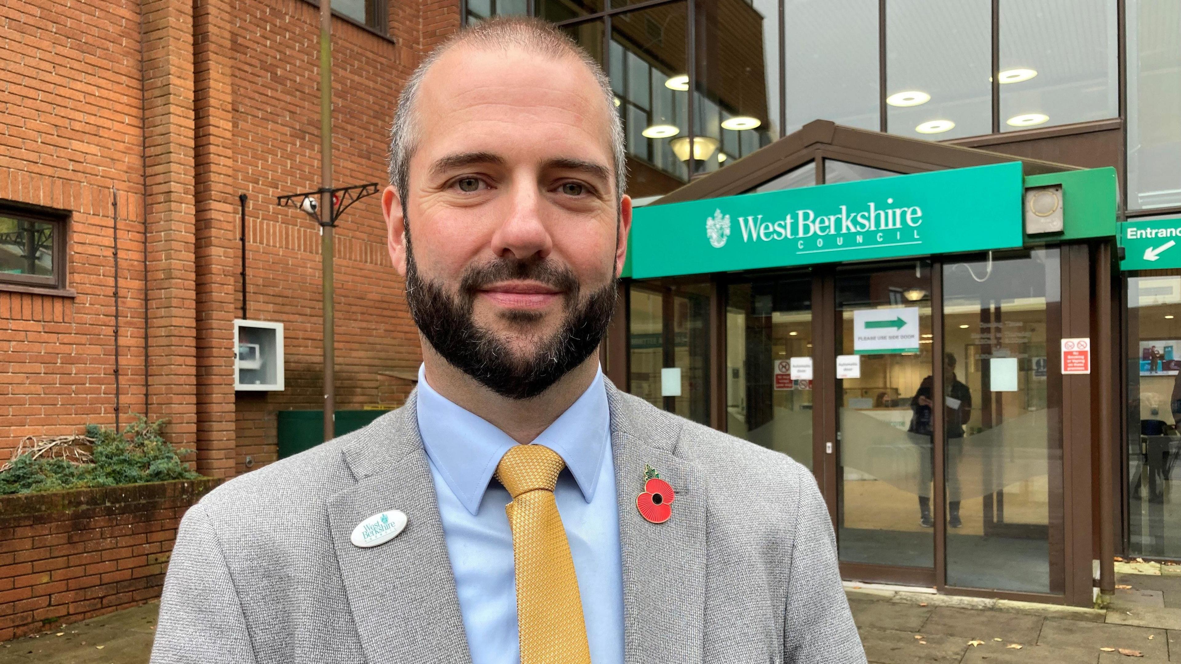 Stuart Gourley smiling at the camera, has a dark beard and is wearing a light blue shirt, with yellow tie and a grey suit jacket with a poppy on his right lapel.