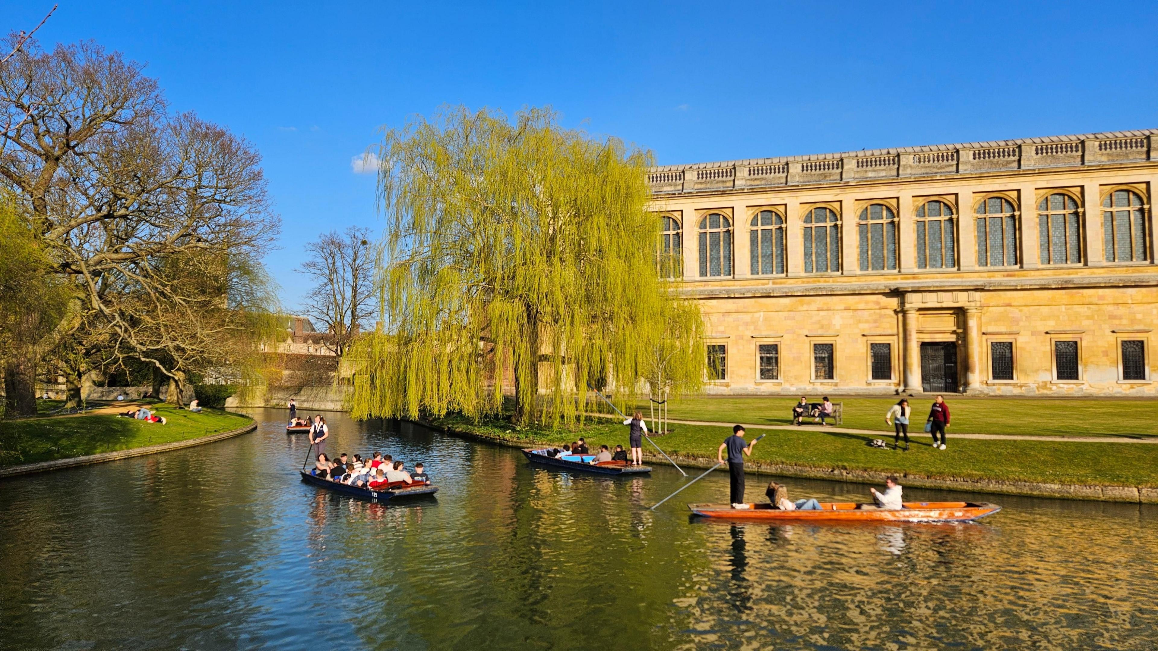 People paddle boats on a river with trees and a building in the background
