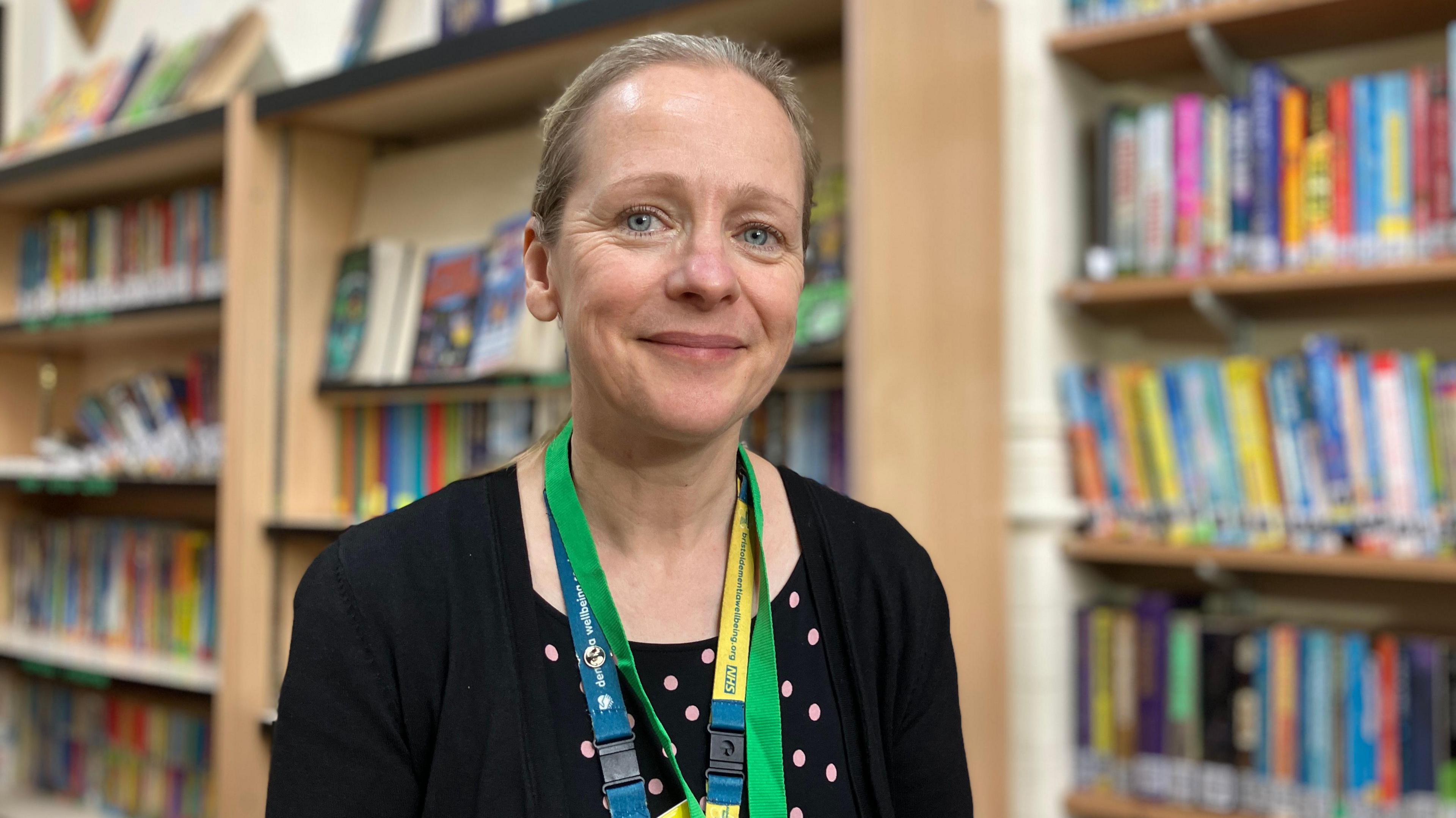 Sally Townsend sitting in Westbury Park Primary School smiling at the camera. She is wearing a black polka dot top underneath a black cardigan, and two lanyards around her neck. She has blonde hair pulled back into a bun and light blue eyes. 