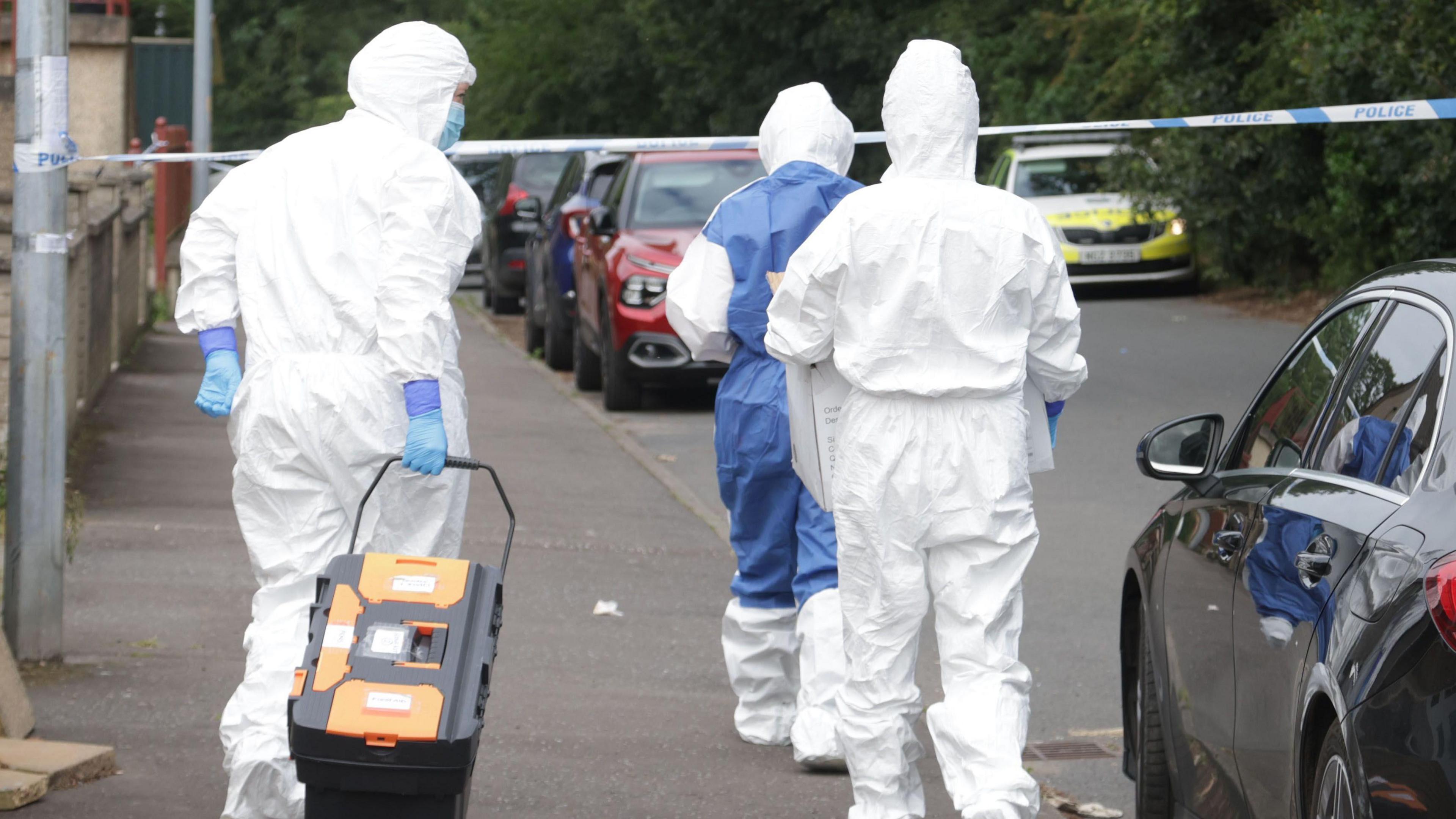 People in white hazmat suits walk down the street with their backs facing the camera. The street has cars parked up and a police car sits in the background.