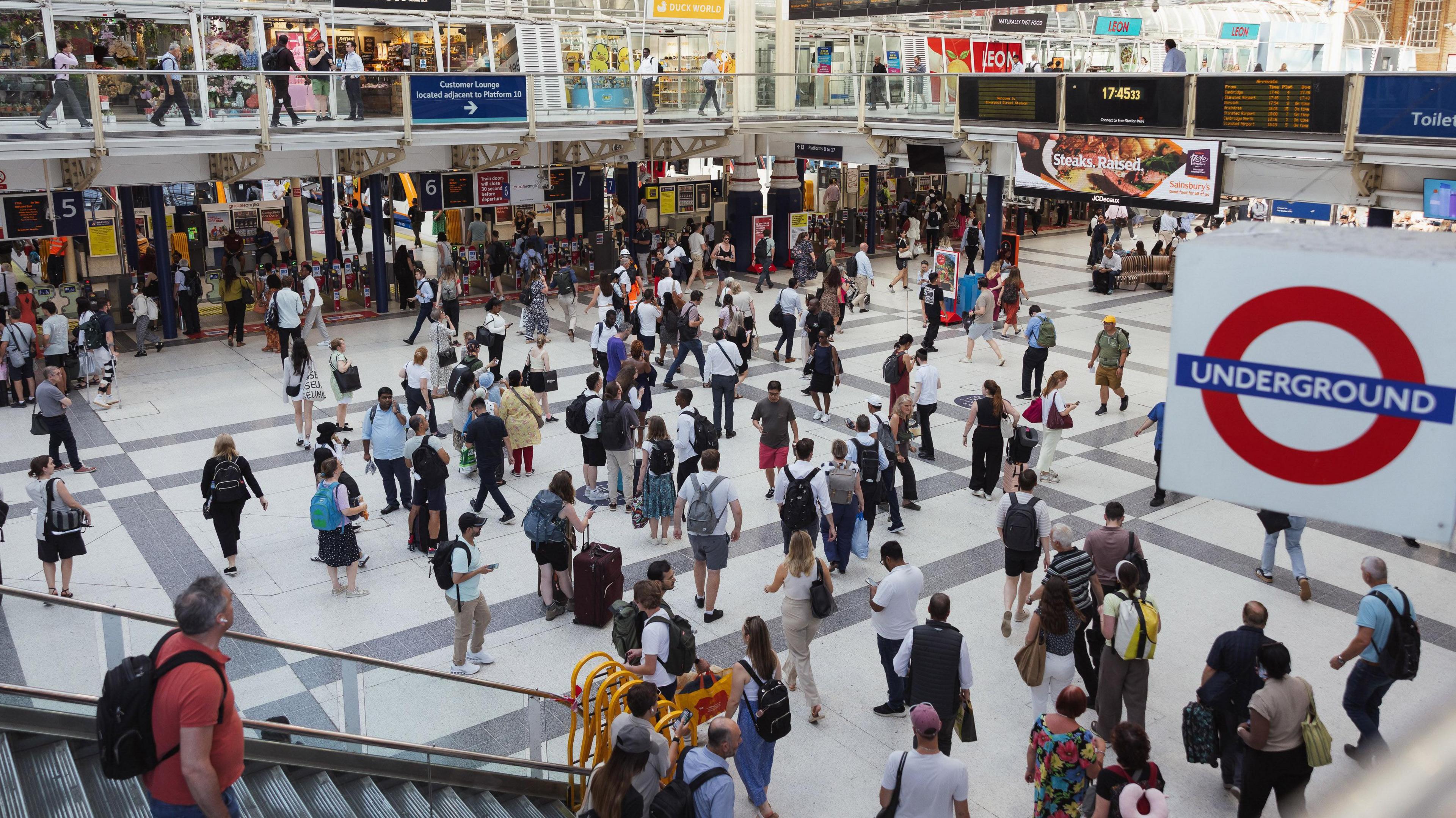 Liverpool Street station concourse at 17:45 shows departure board and Tube roundel to the right of passengers surrounded by shops and platforms 5-7