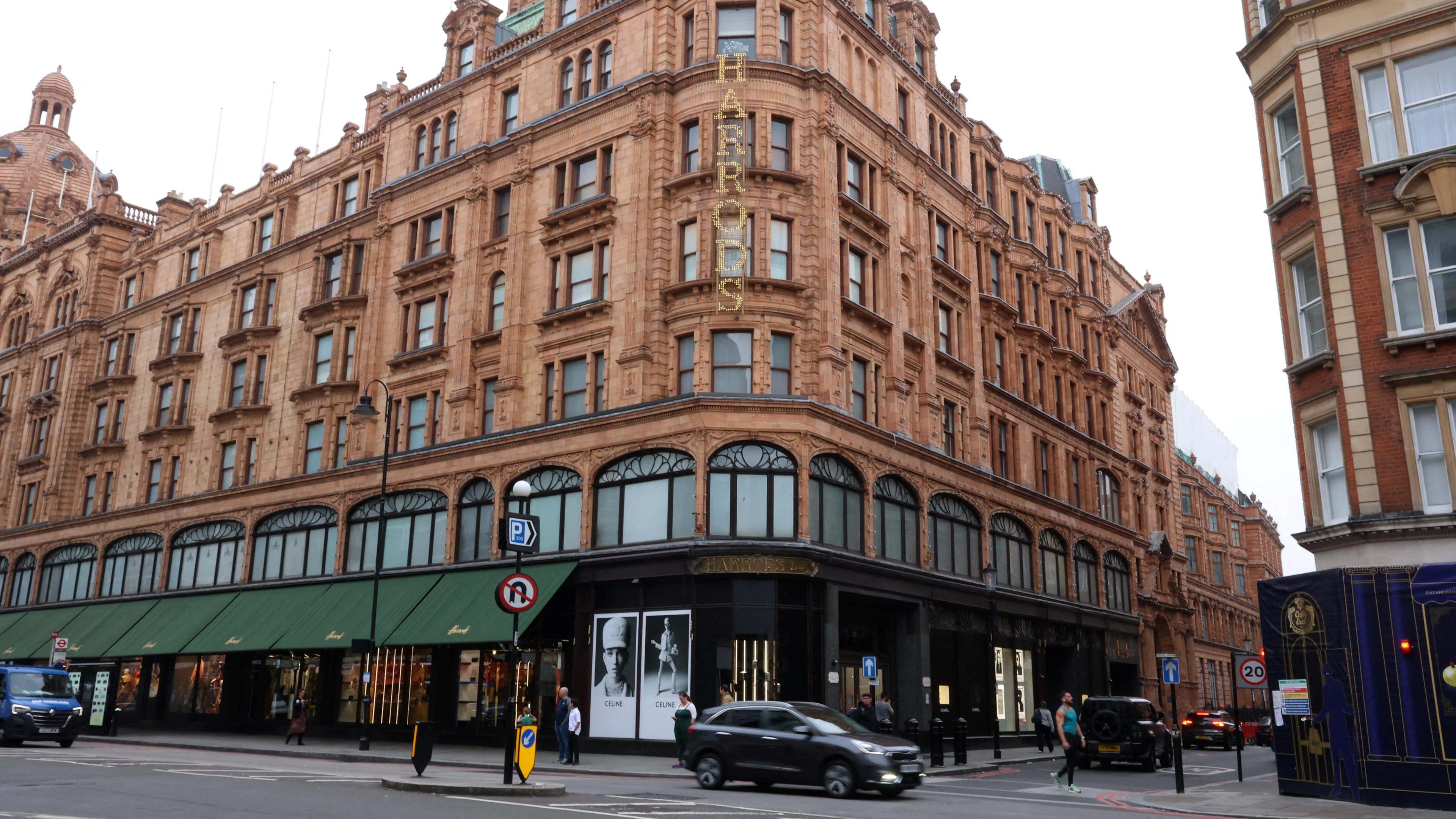 An exterior of Harrods department store. Traffic drives in front of the building.