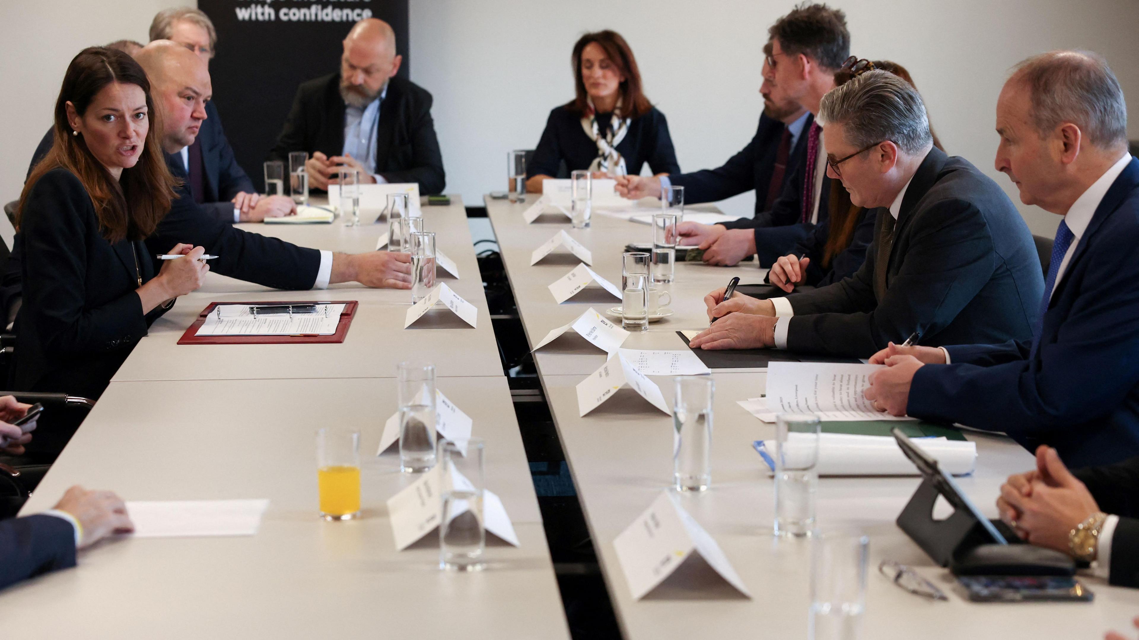 Ministers sit round a white table. They wear suits. There are name cards, paper, clipboards and glasses of water on the table.