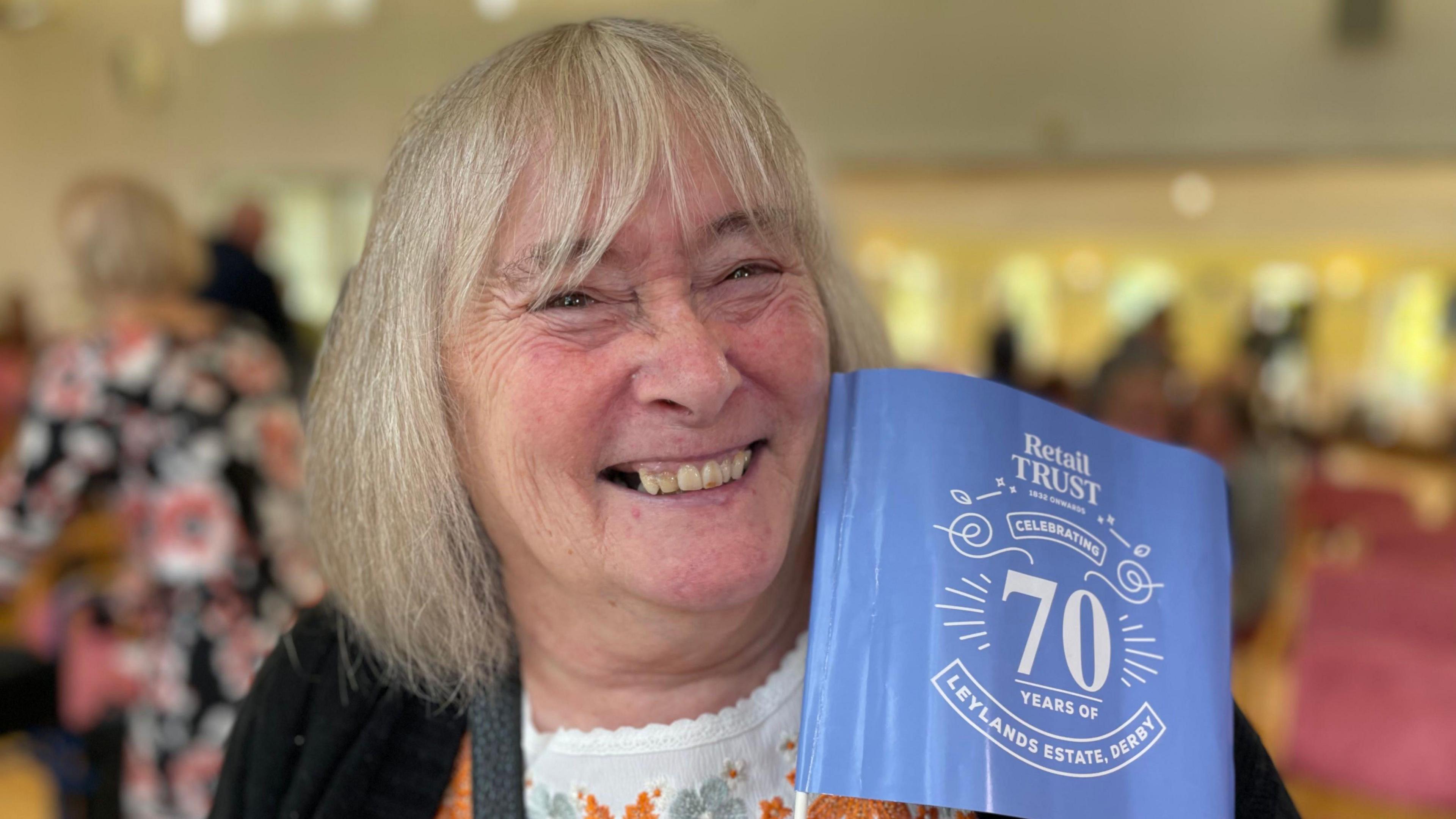Josie Gadsby, an older women with a short greying bob haircut. She is smiling and holding a small blue flag commemorating 70 years since the opening of a retirement complex in Derby.