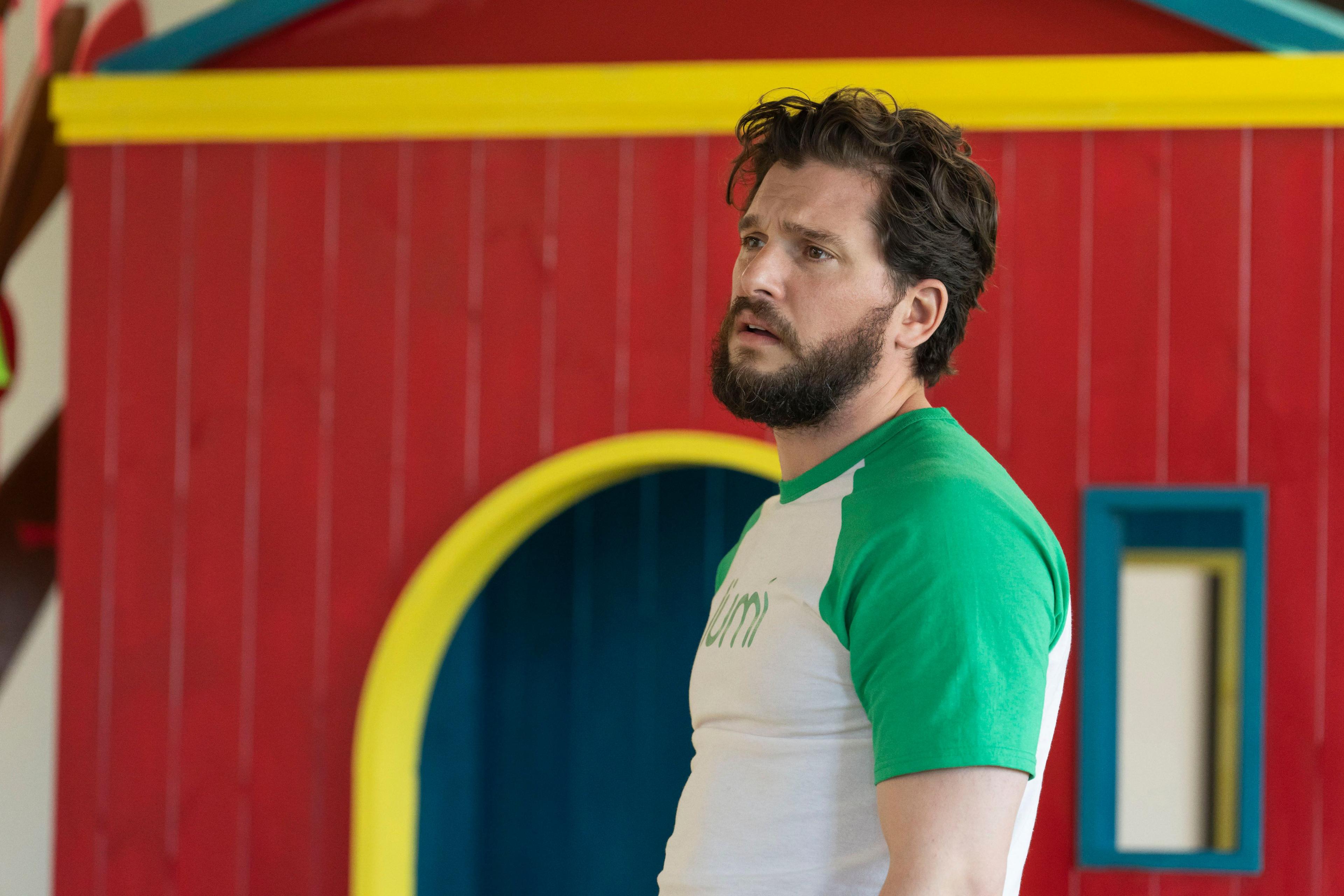 A man, frowning, with brown hair is dressed in a white short-sleeved t-shirt with green detailing