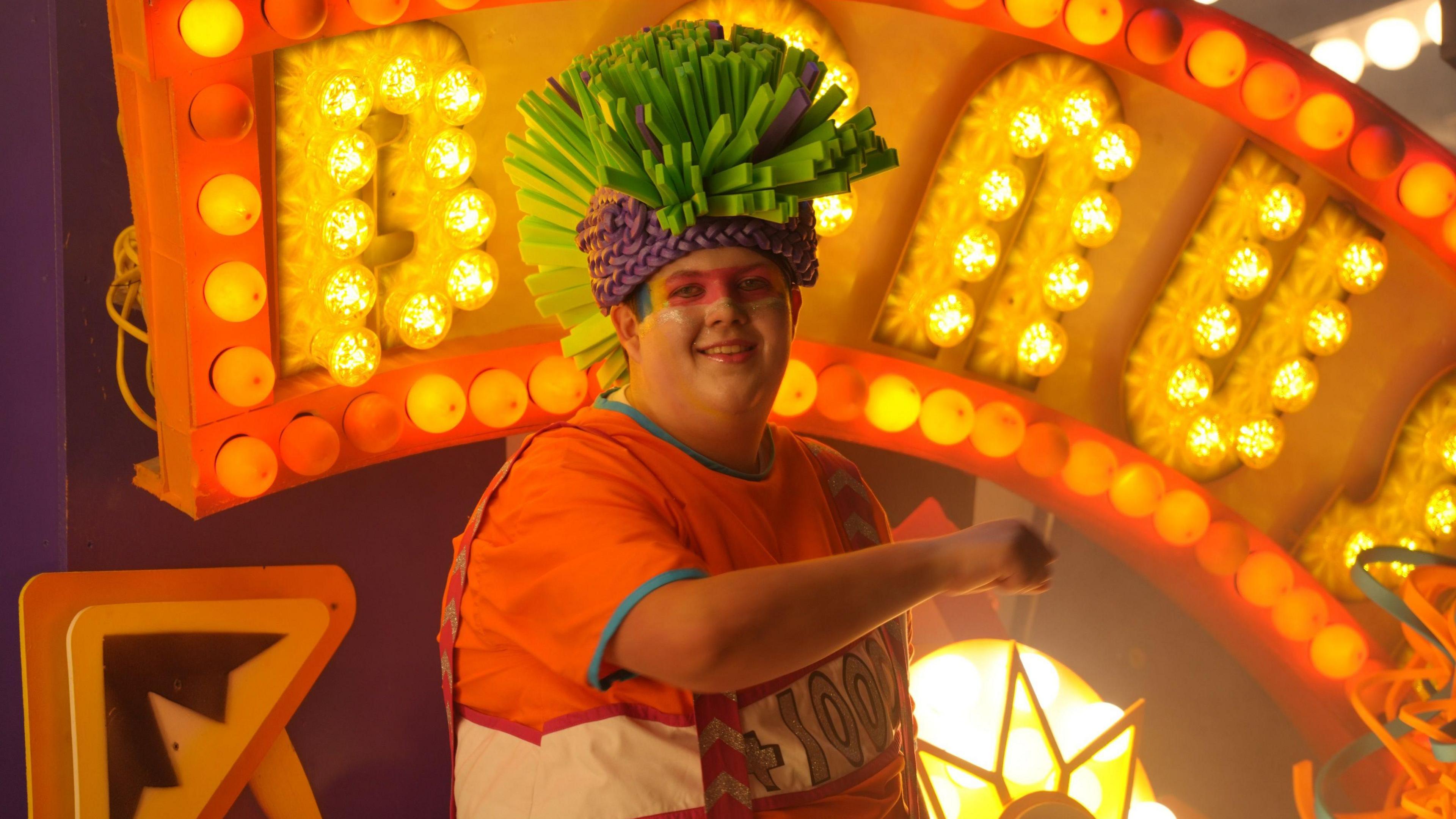 A man wearing a bright and spiky green and purple hat dances on top of a cart