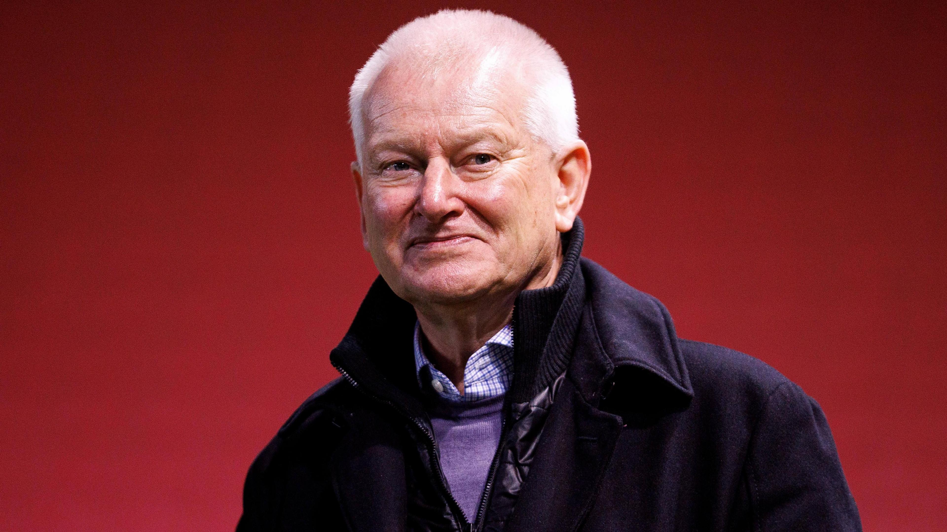Bristol City owner Steve Lansdown wearing a black coat and purple jumper over a purple chequered shirt with a red background behind him