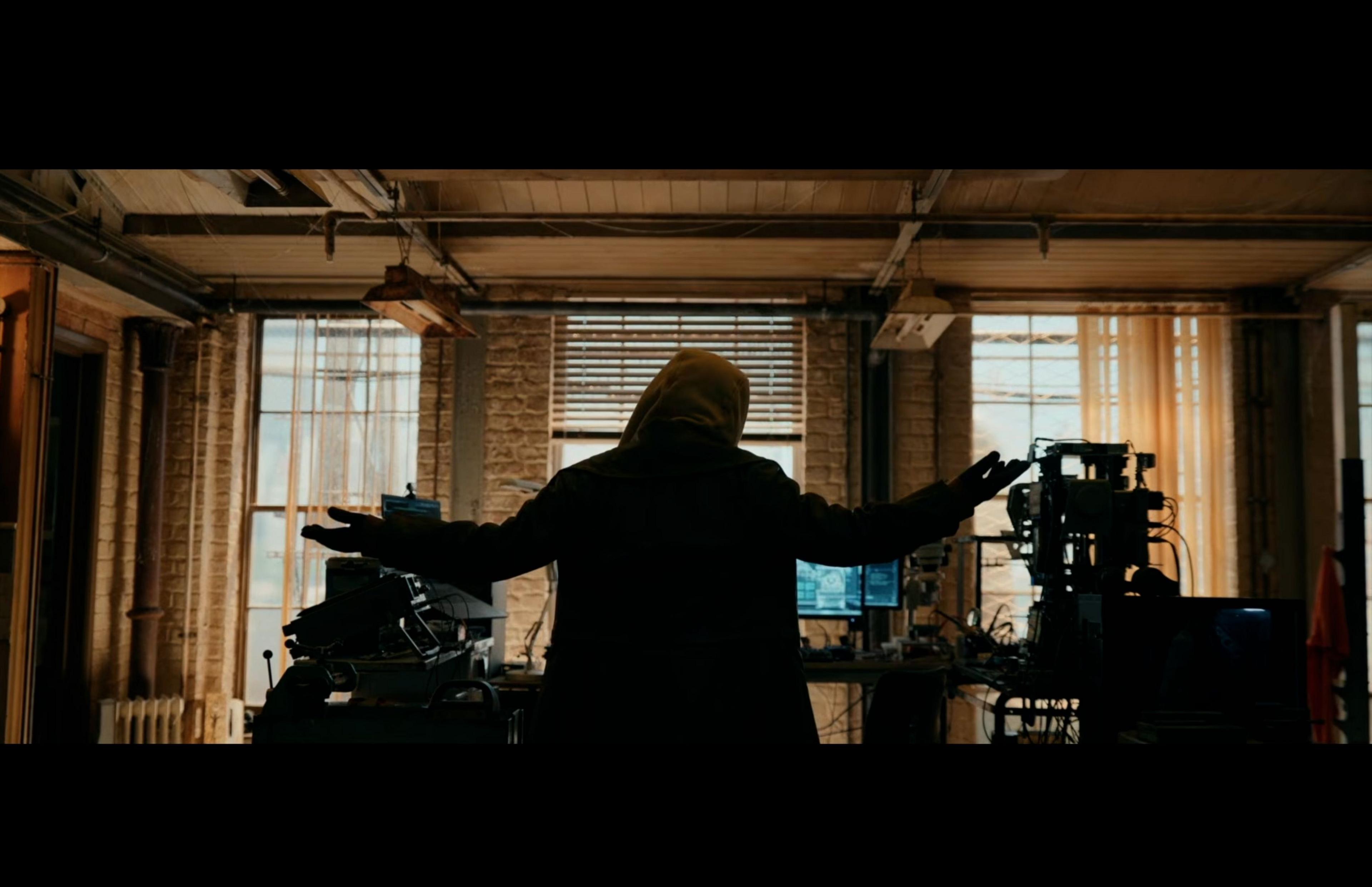 A hooded figure stood in front of computer monitors raises their hands. 