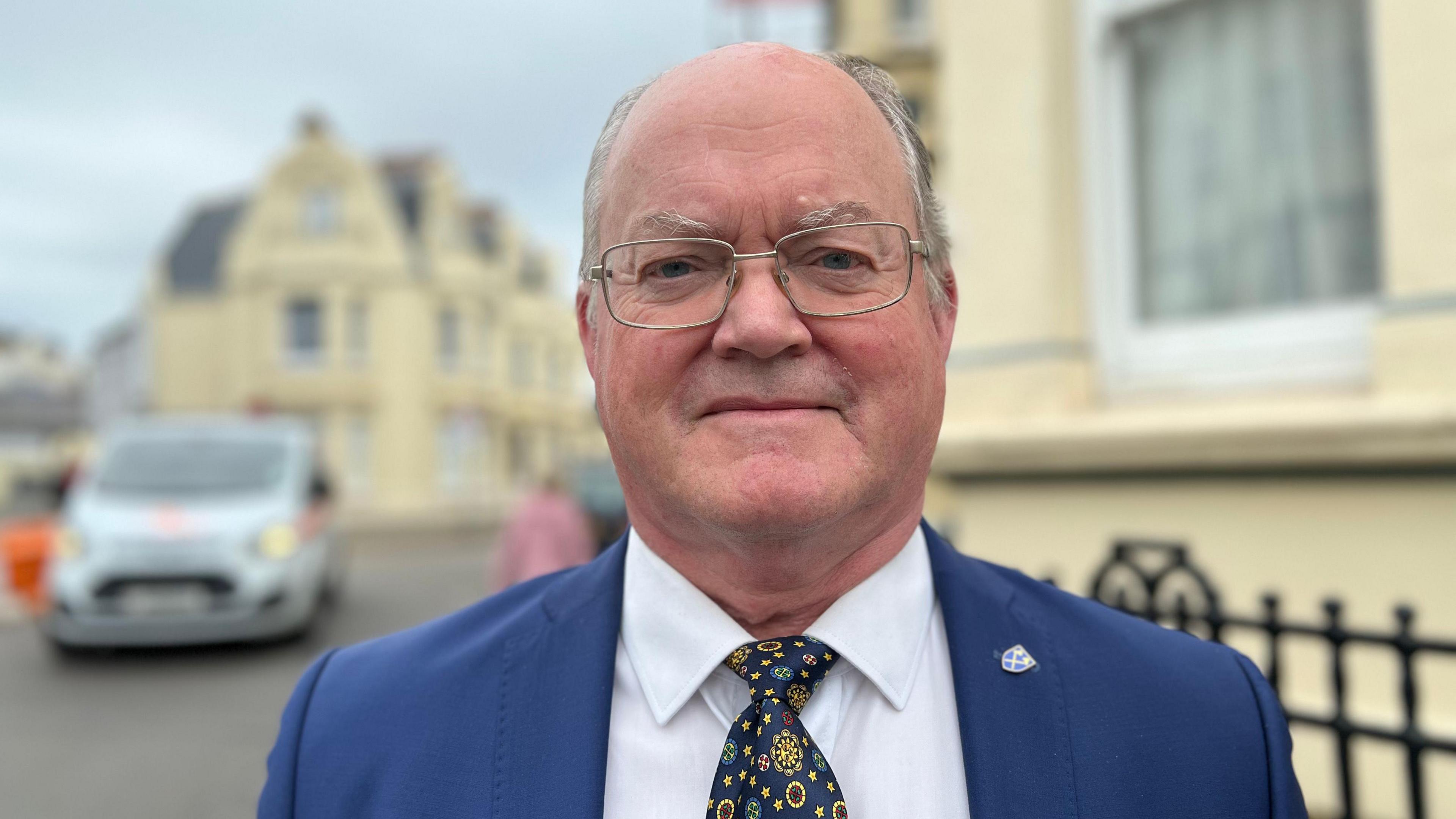 A bald man with grey hair on the sides, wearing a blue suit, white shirt and patterned tie. He wears glasses and is standing in a residential street.