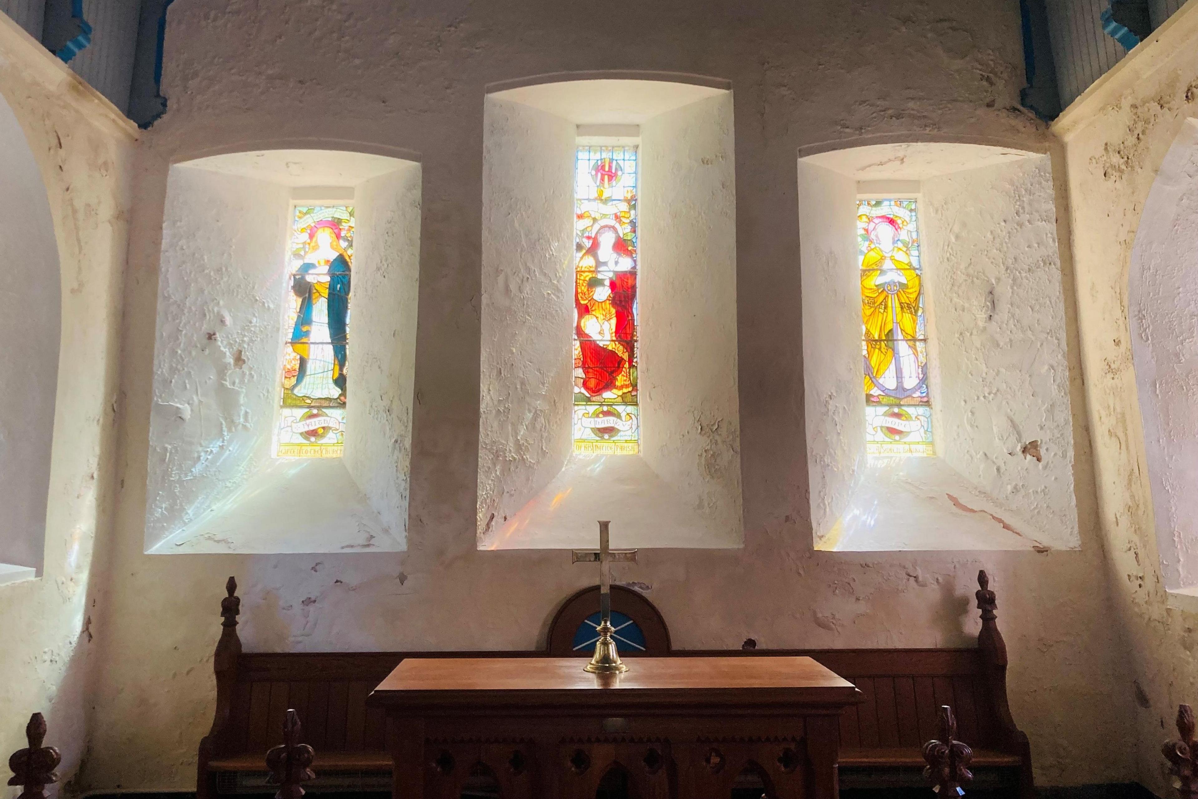 Three narrow stained glass windows in a church with light streaming through them.