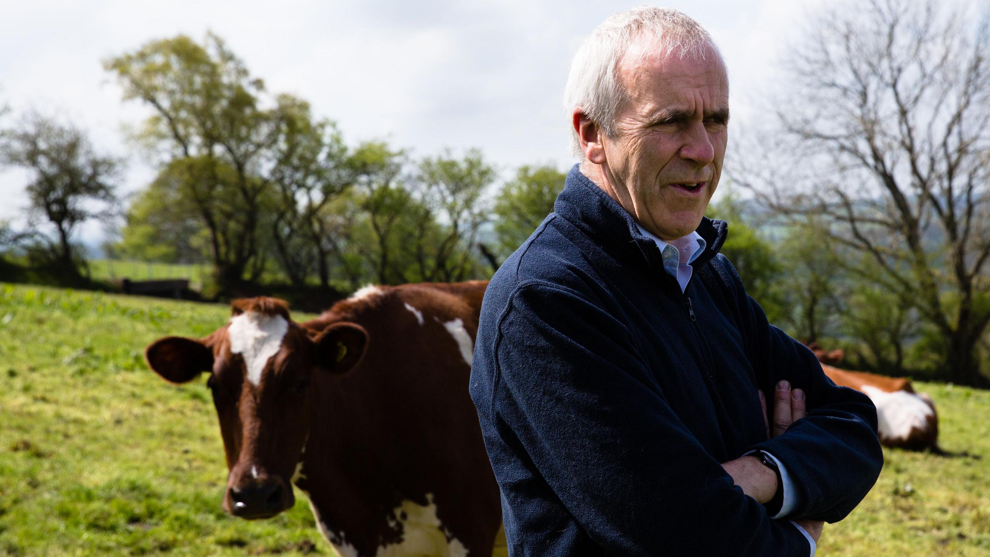 Patrick Holden - a middle aged man with thinning grey hair - standing in a field in front of a cow 