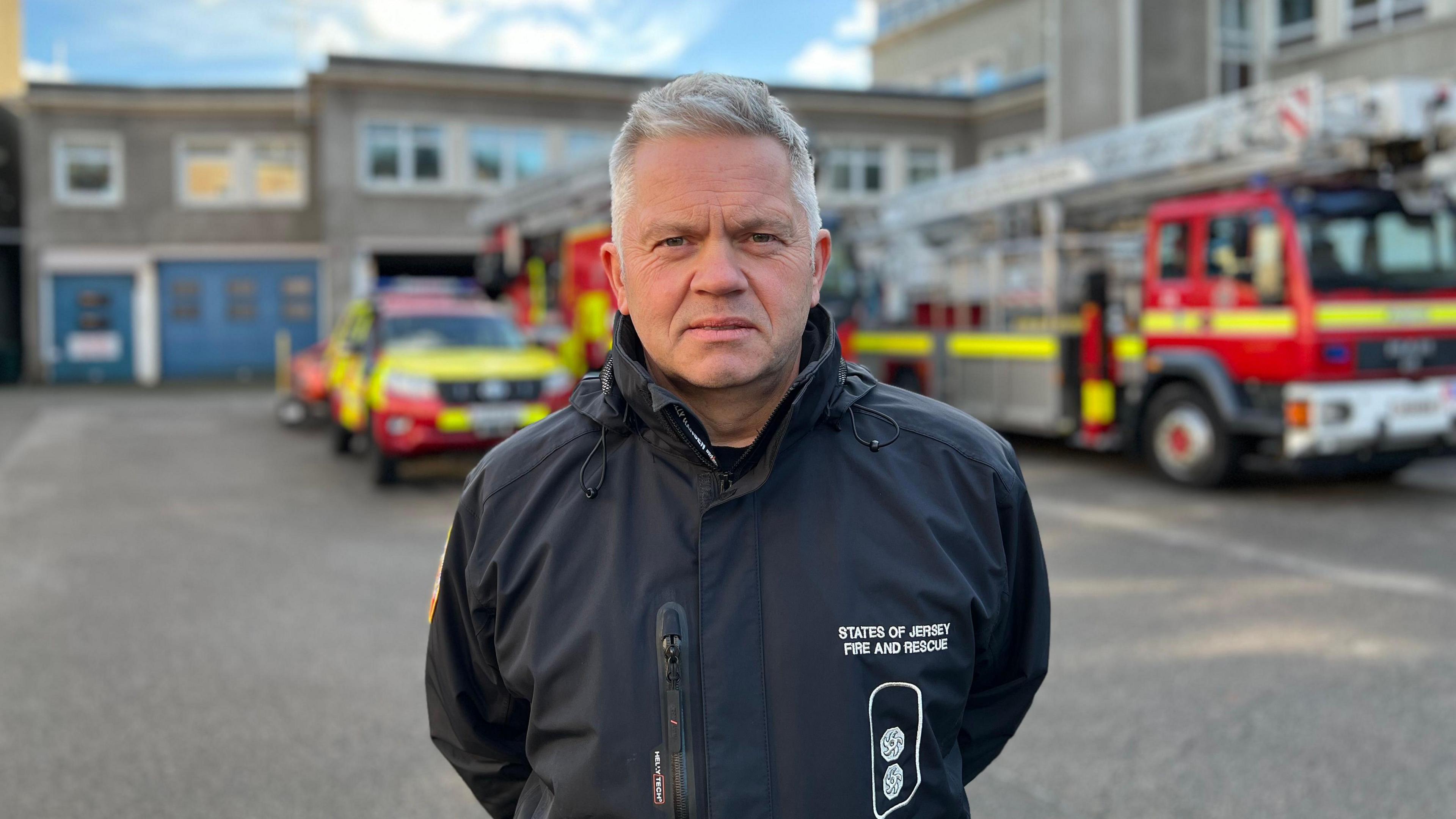 A man wearing a black coat with the States of Jersey Fire and Rescue logo on it, behind him are parked fire vehicles including two fire engines.