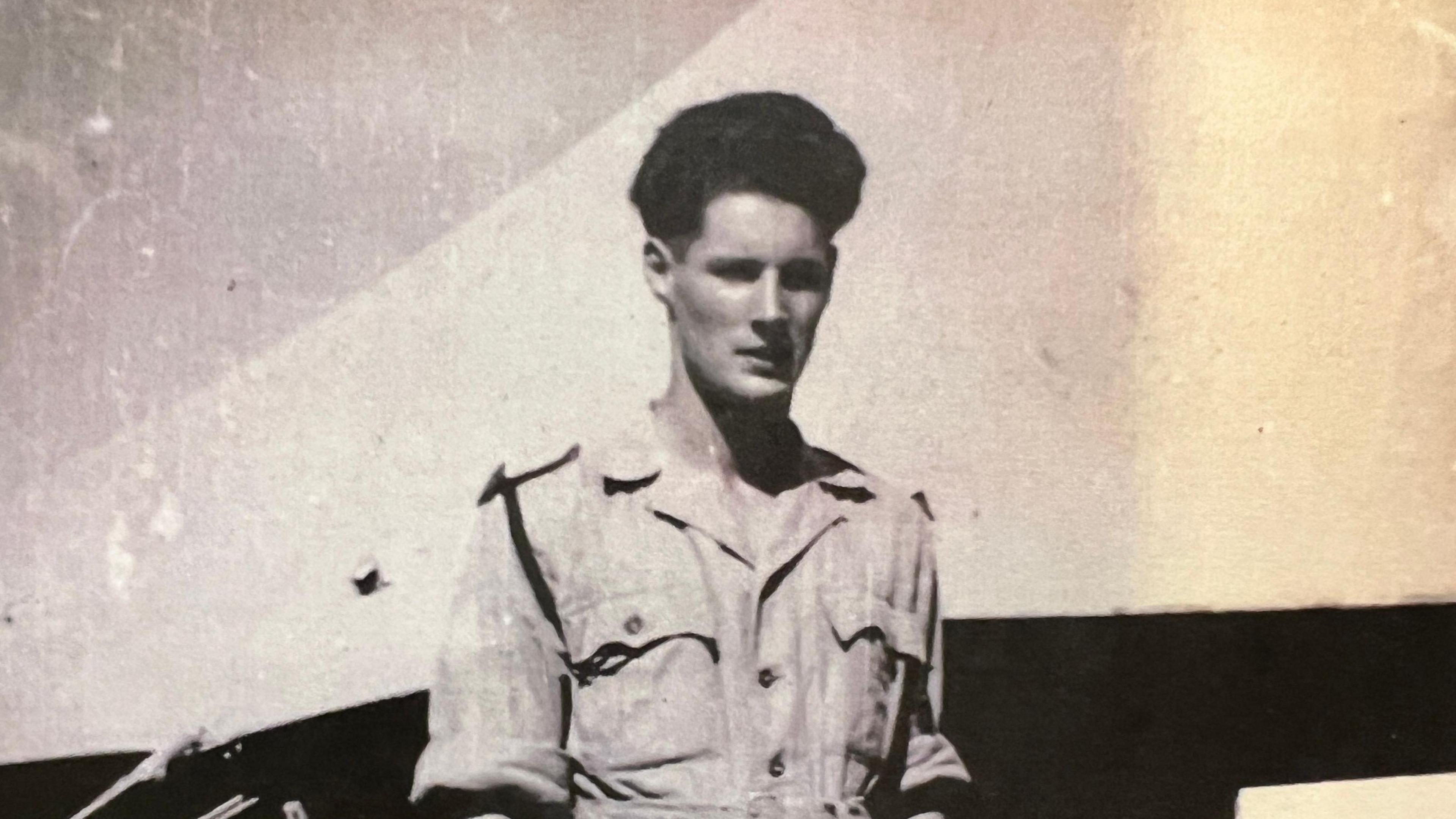Black and white photograph of a person standing outdoors in a short-sleeved military-style uniform with buttoned pockets and a waist belt.