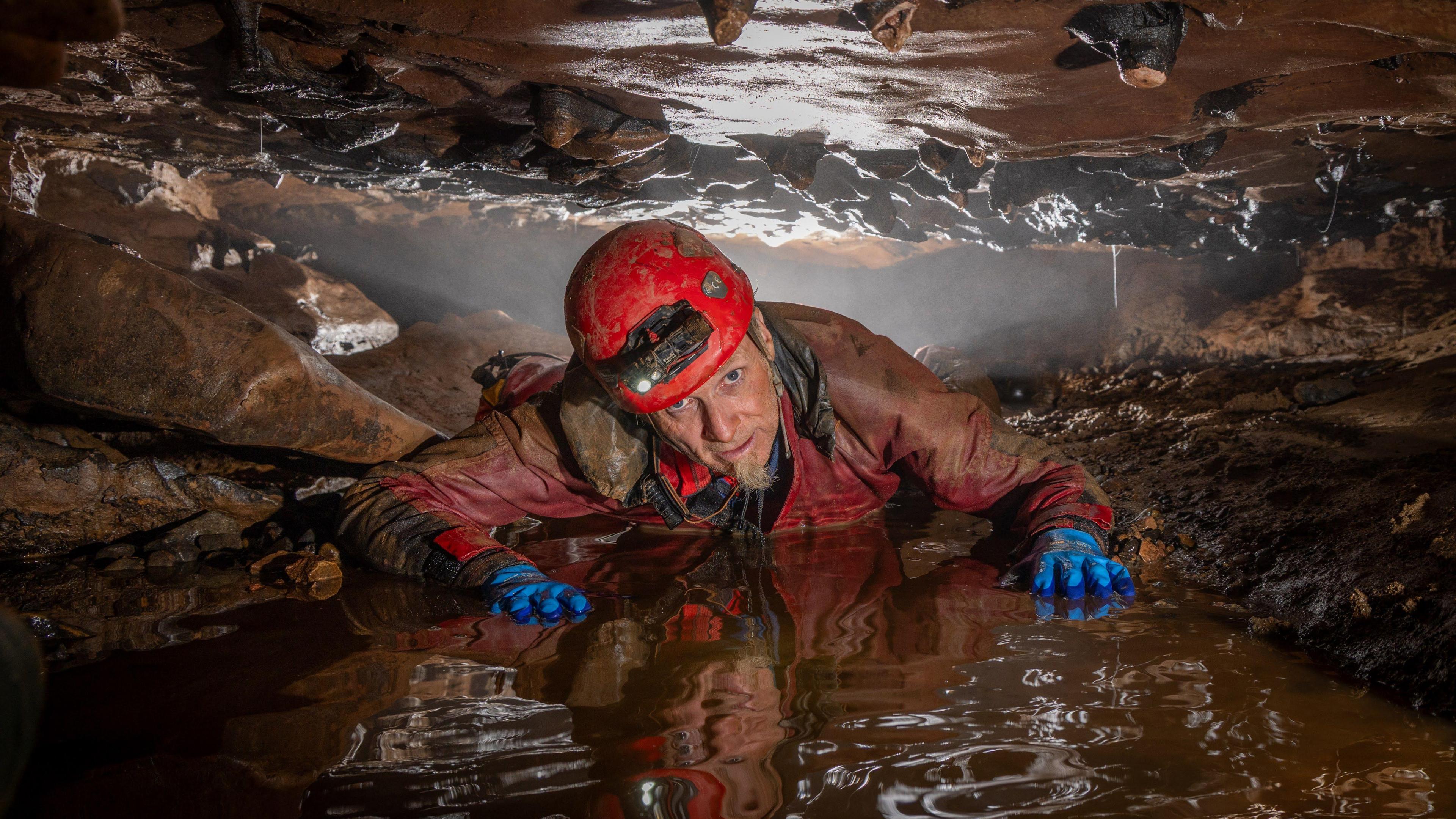 A caver from the exploratory team wearing waterproofs, blue gloves and a red helmet with a light on on his front between a small gap of rock that sits above and below him. He has his hands on the wet ground in front of him and is looking at the camera.