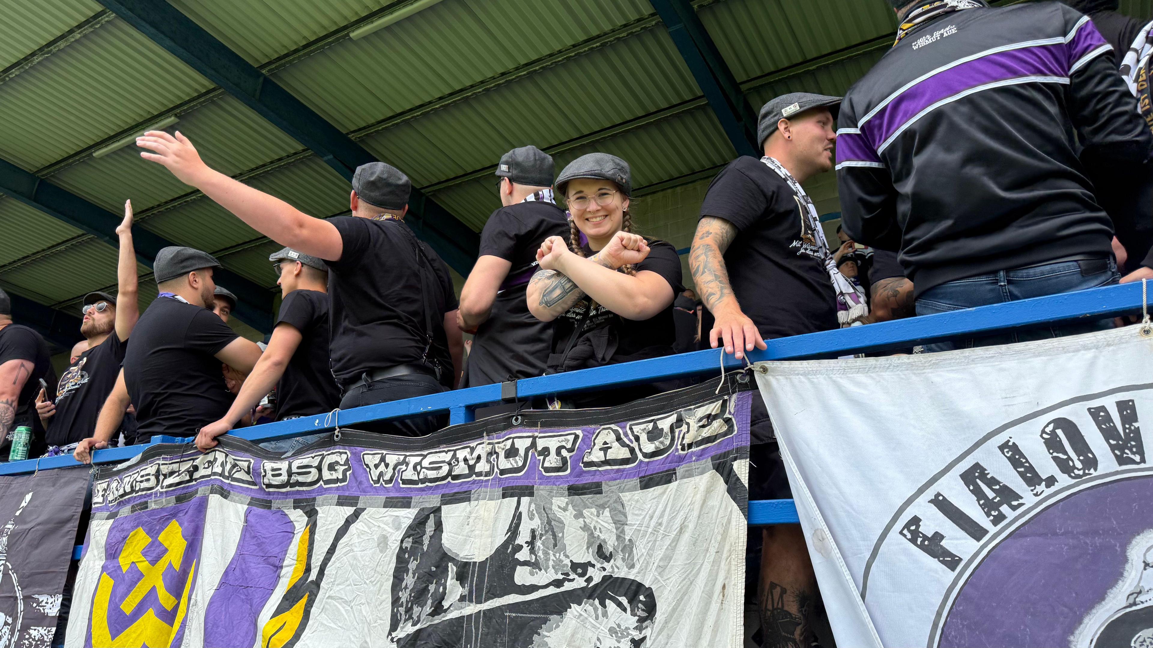 A line of German fans, dressed in black. One fan is making an 'X' sign with her arms. They are all wearing grey flat caps. There are flags of the German team tied to the stand. 