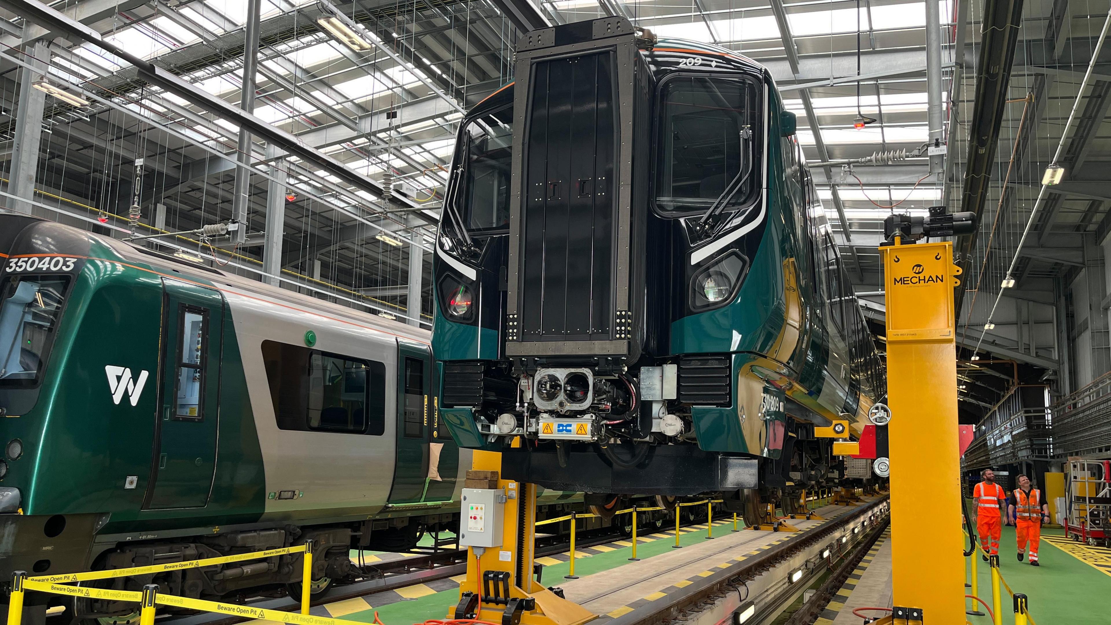 Two trains in a depot. One has been lifted off the ground. Two station workers in orange high-visibility clothes are walking beside the train.