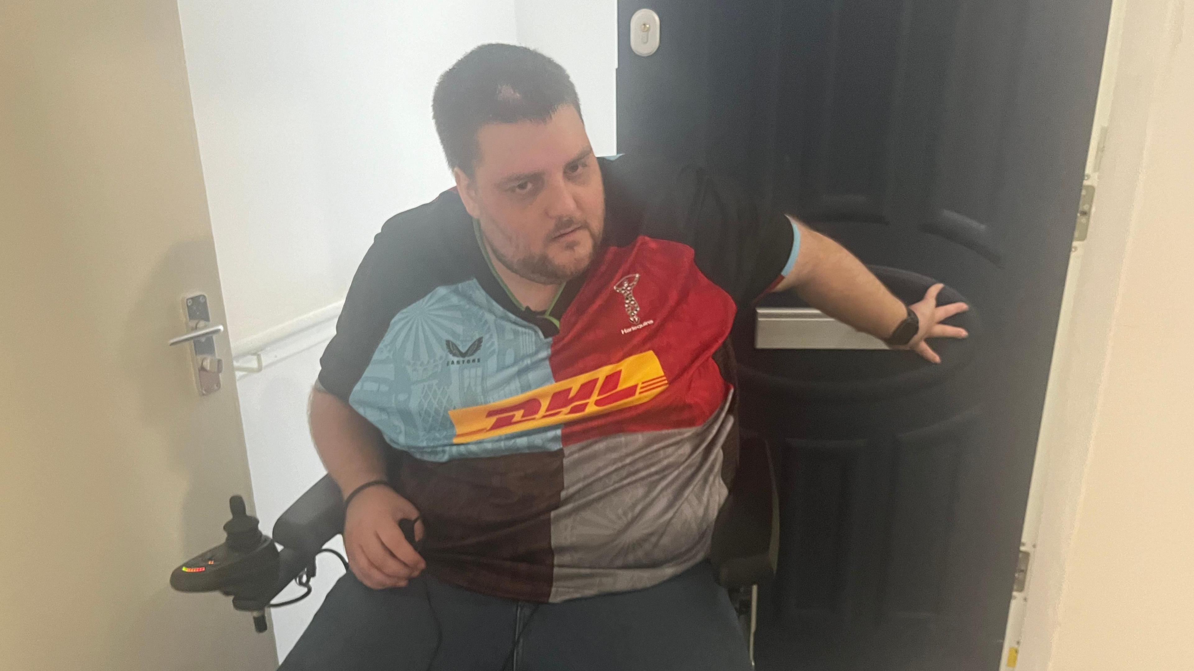 Nathan Rollinson, a man in a red and blue rugby shirt, trying to reach behind him to wedge open the front door while in his wheelchair