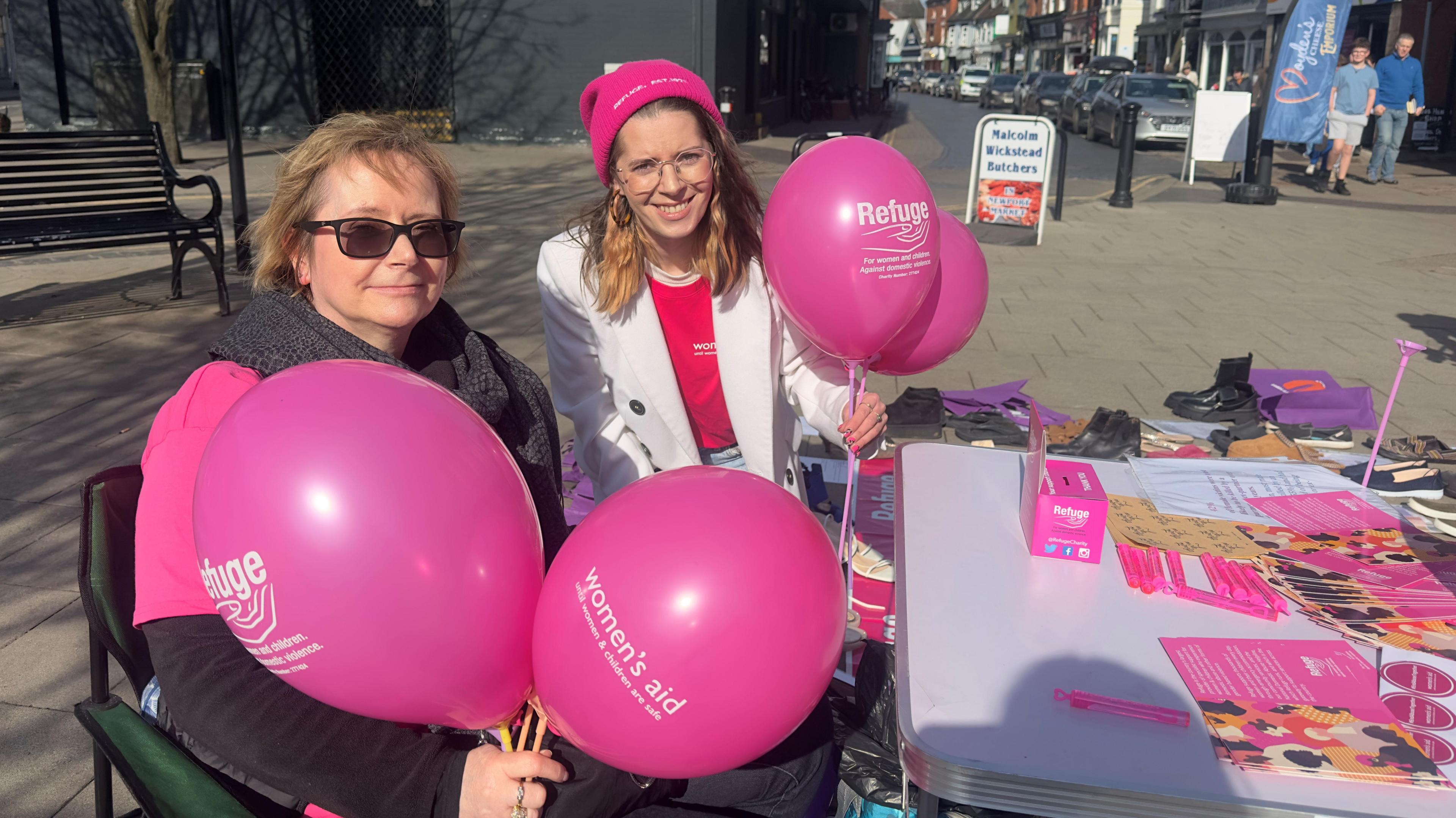 Two women are sat at a table in a high street. They are holding Women's Aid and Refuge branded balloons. On the table are leaflets and other promotional literature. In the distance are pedestrians and a row of parked cars.