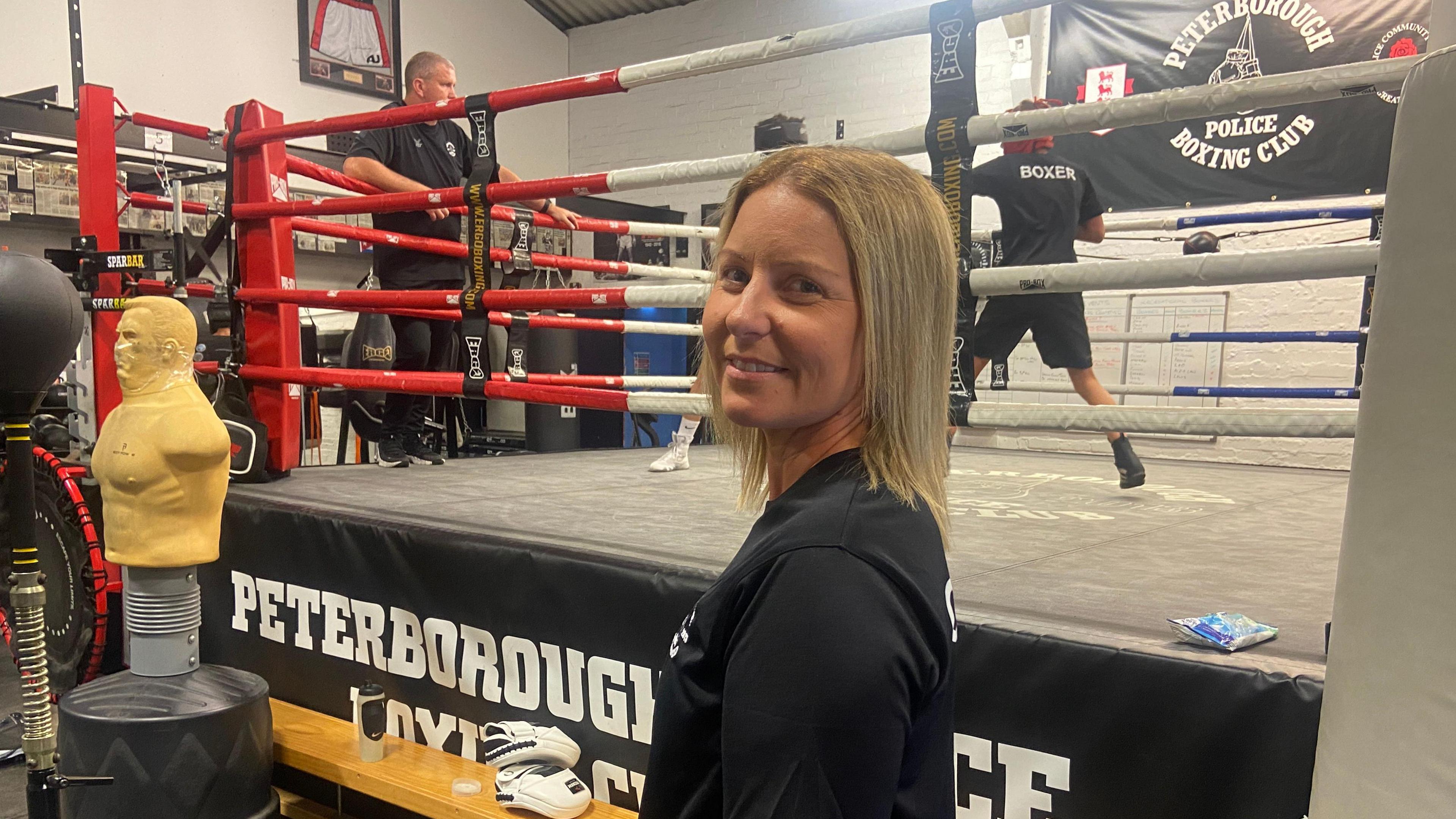 Vicki Baker stood in front of a boxing ring, which has one person inside training and another watching from outside. She is side on to the camera and has her head turned to look at the lens. Vicki has shoulder-length blonde hair and is wearing a black t-shirt. 