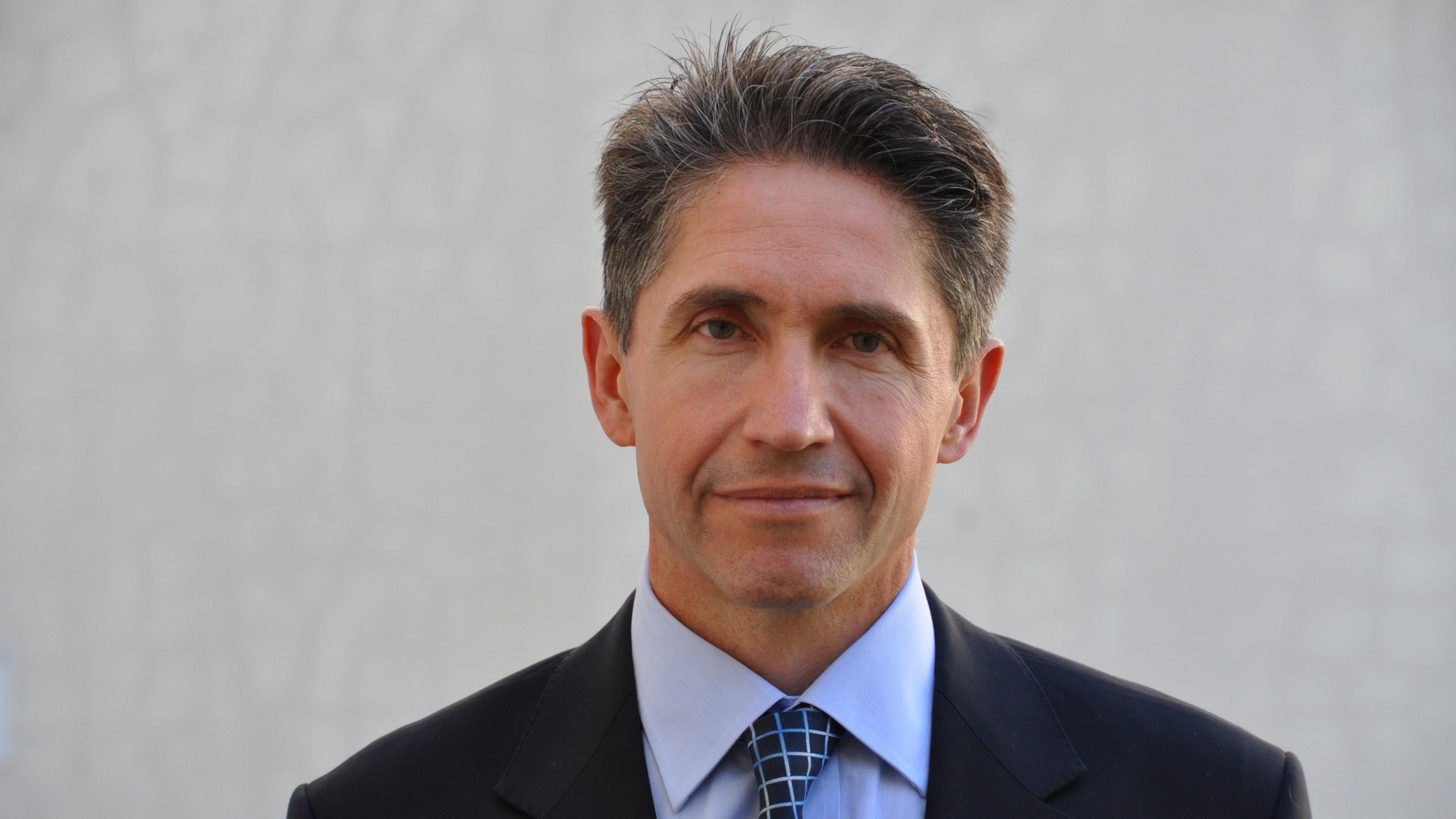 BBC's Paul Barltrop wearing a blue shirt and black suit. He is smiling while stood in front of a white wall.