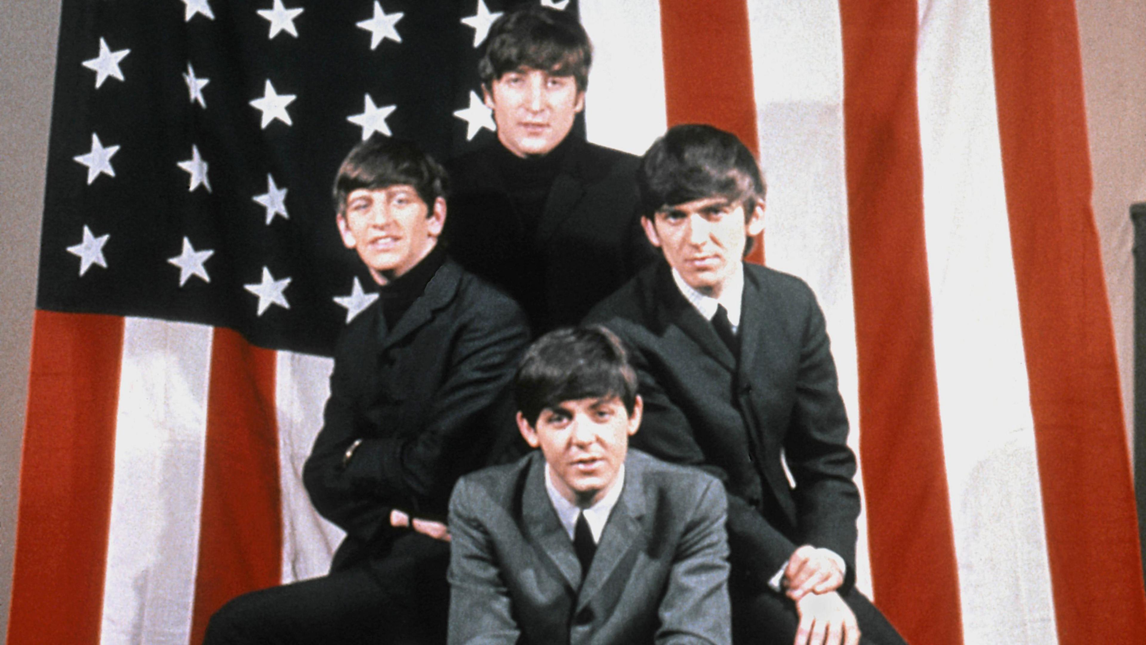 The Beatles, in black and grey suits, shirts and polo neck jumpers, pose for a portrait in front of an American Flag