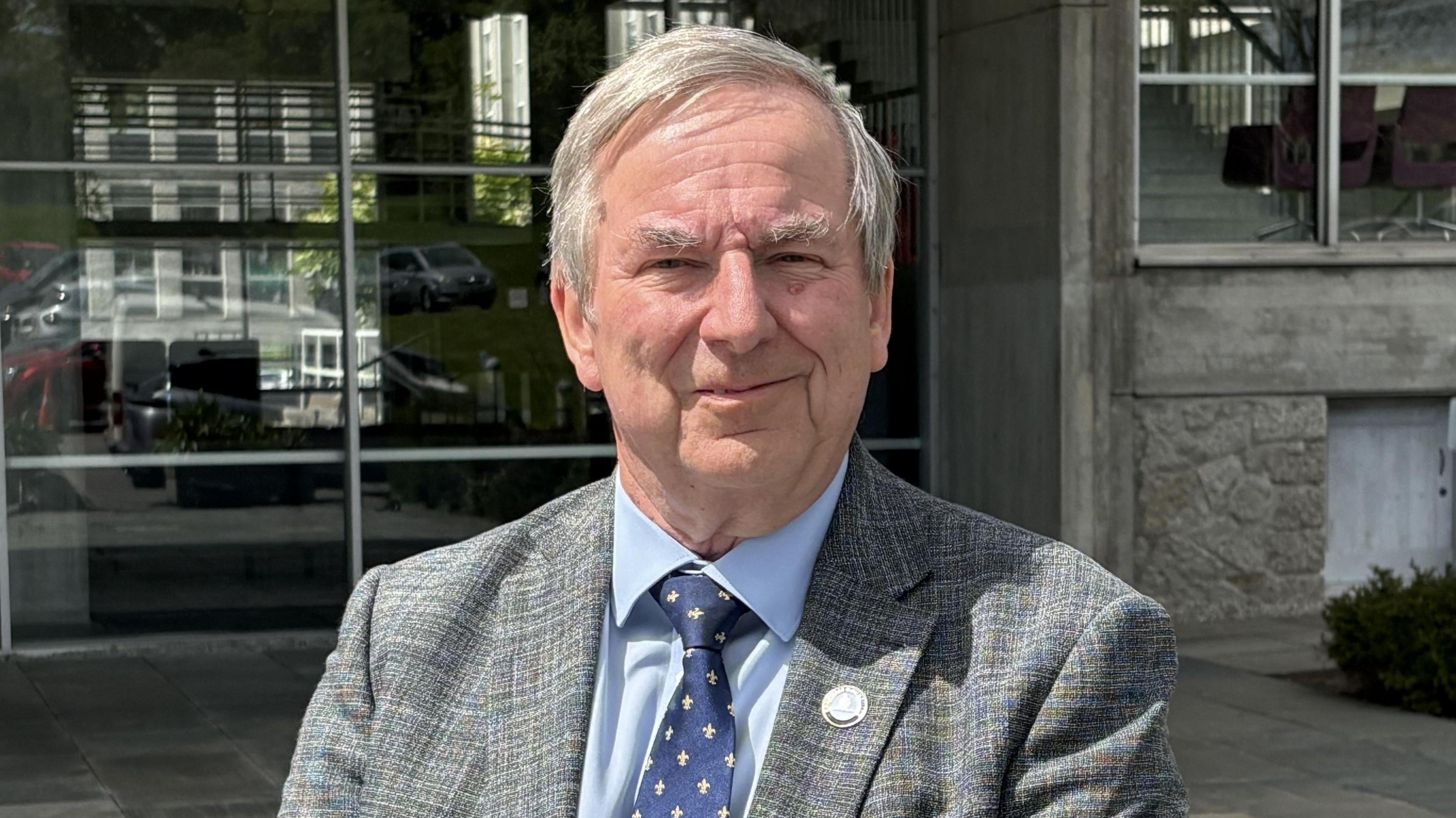A man with grey hair in a grey jacket with a blue shirt and dark blue tie standing in front of County Hall in Truro