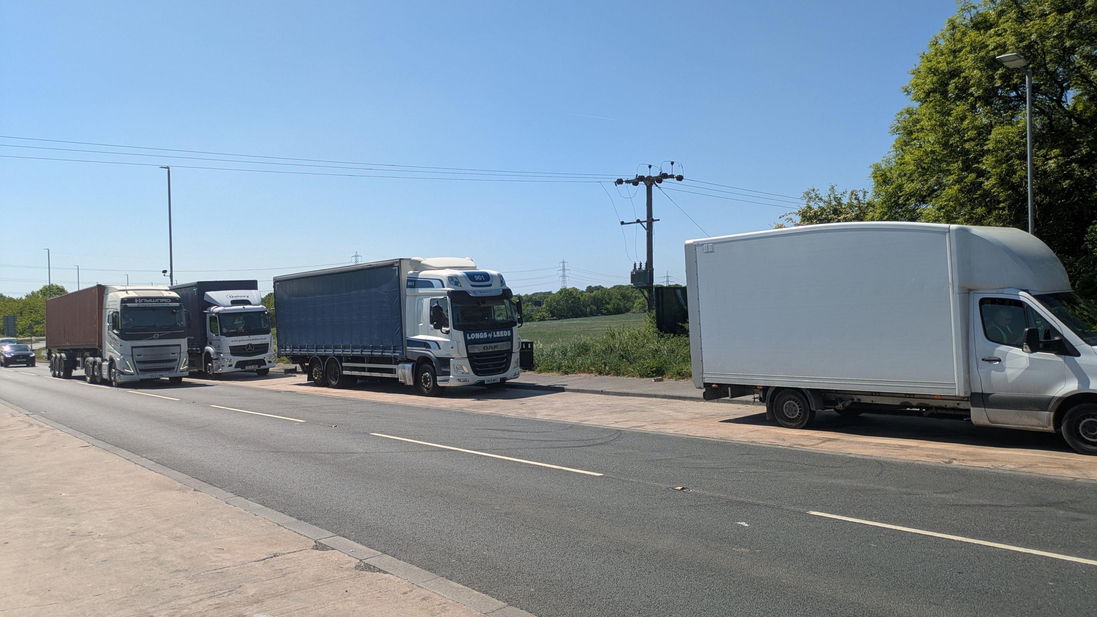A row of lorries parked up in a lay-by on a sunny day. Another lorry is seen driving past.