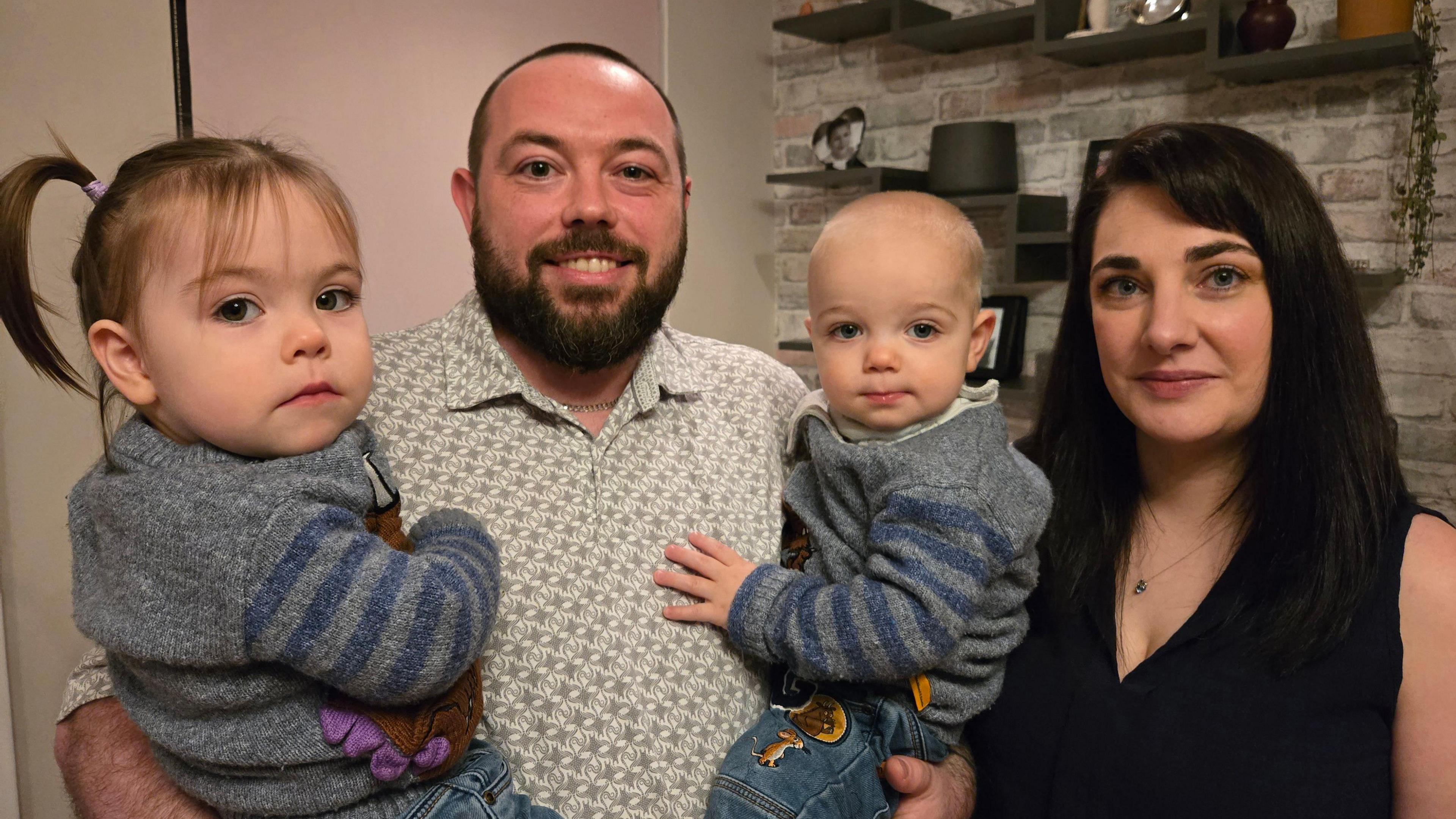 Two adults and two young children. The dad is holding both children, who are wearing matching gray sweaters with blue striped sleeves and jeans featuring colourful patches. The children are each holding a small plush toy. The background shows a light-coloured brick wall with shelves displaying decorative items such as framed photos, a black hat, and small plants
