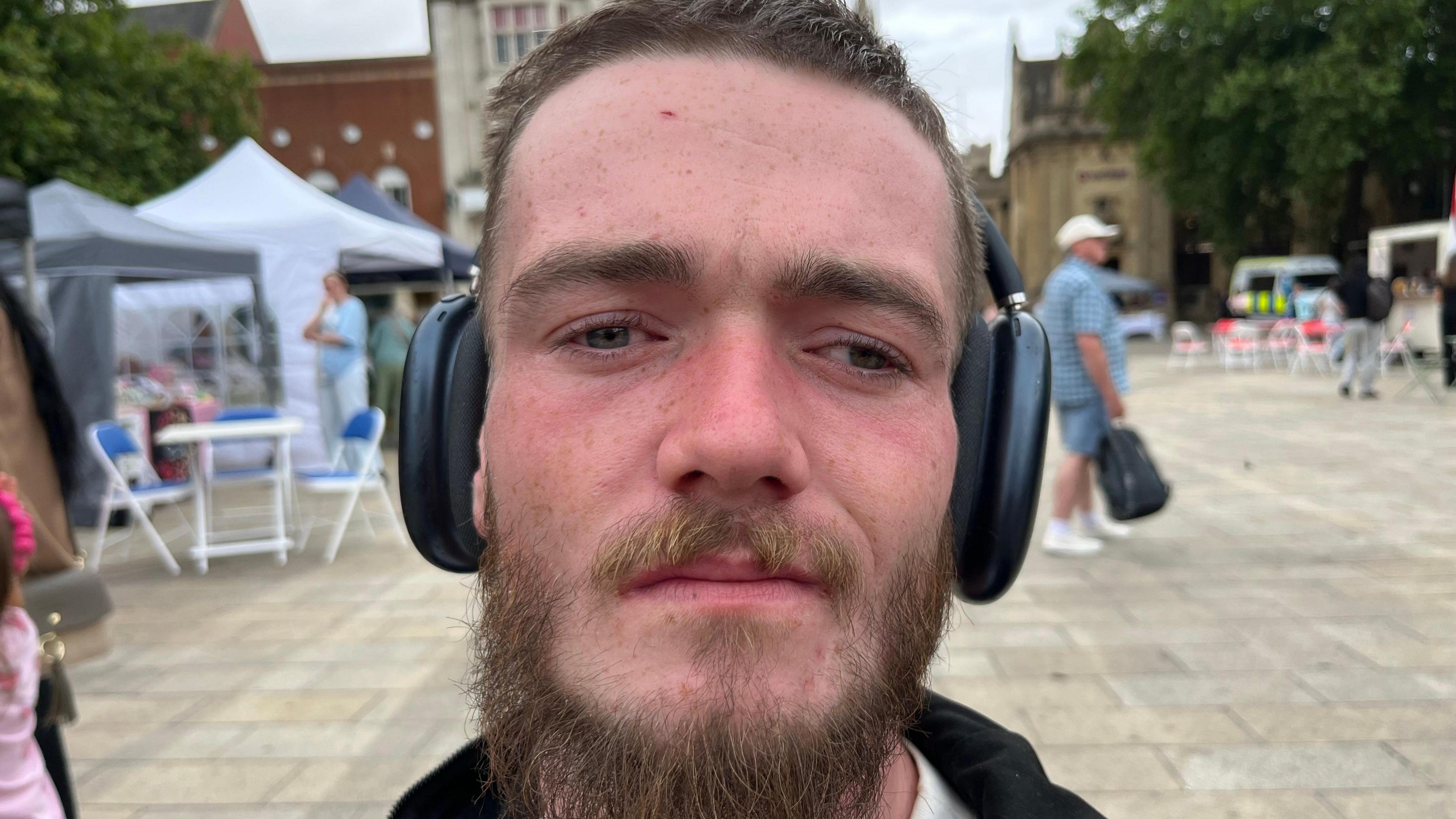 Daniel Slack stands in a square with dark hair, a beard and a mustache. He is wearing large black headphones. There are chairs and gazebos behind him.