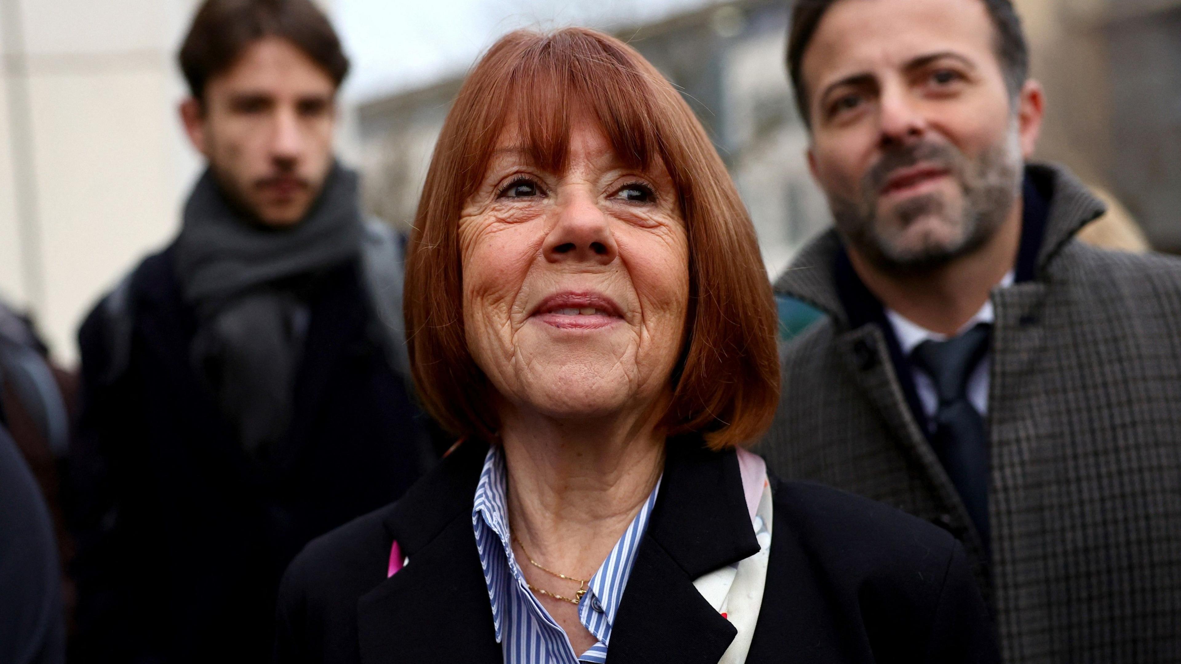 Gisèle Pelicot, wearing a black jacket and striped blue and white shirt, smiles as sge arrives with her lawyer at a courthouse