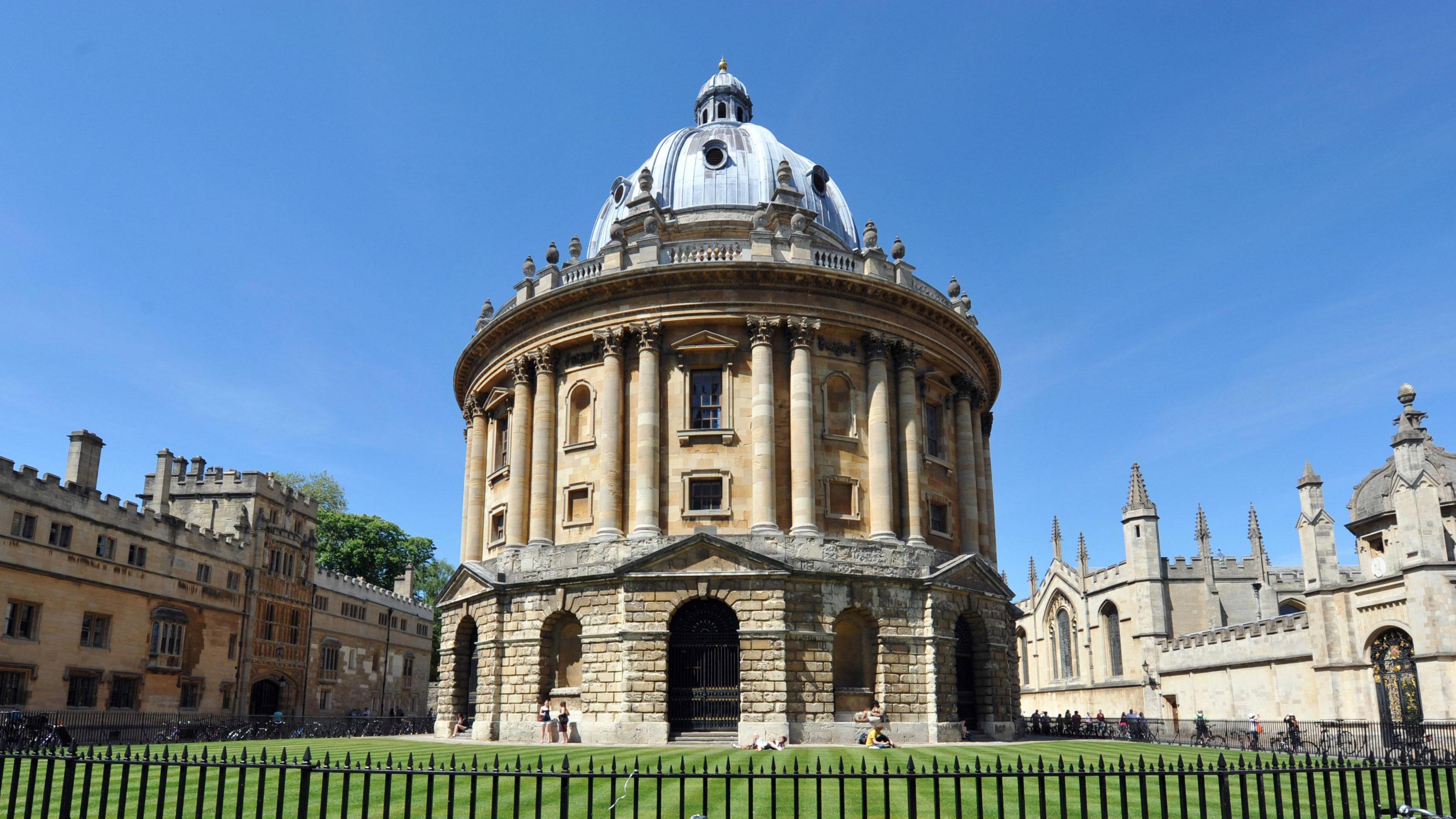 Oxford's Radcliffe Camera building a sunny day - with blue skies above and green grass in the foreground. The building is dome-shaped and made from light-coloured stone. There are several other buildings in the background made from the same stone.