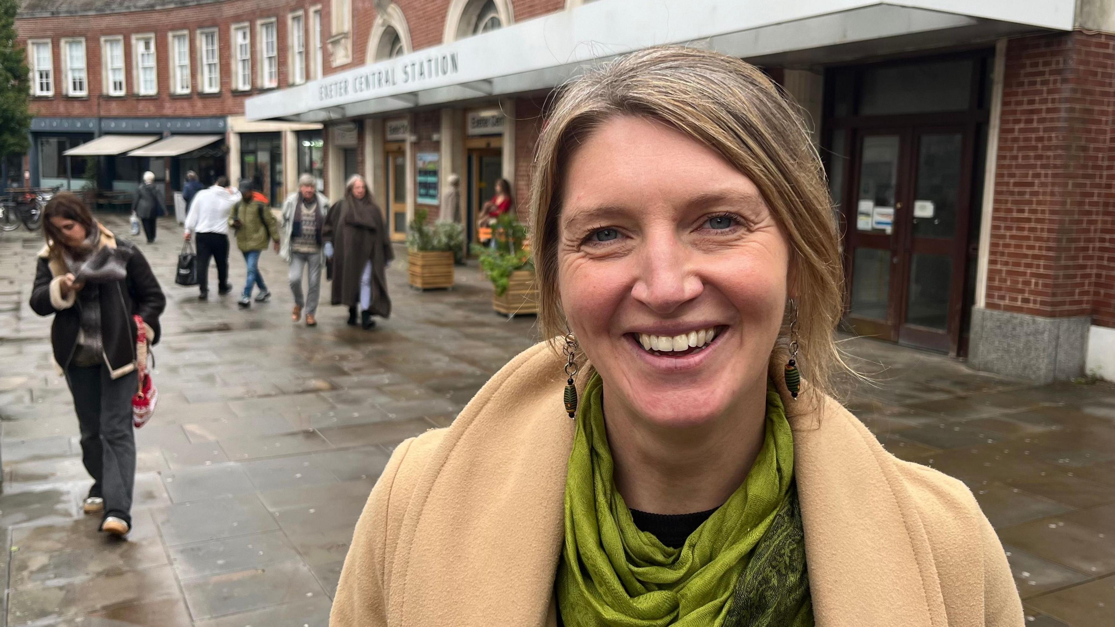 Nicola Wheeler wearing a green scarf and a beige coat in front of Exeter Central railway station