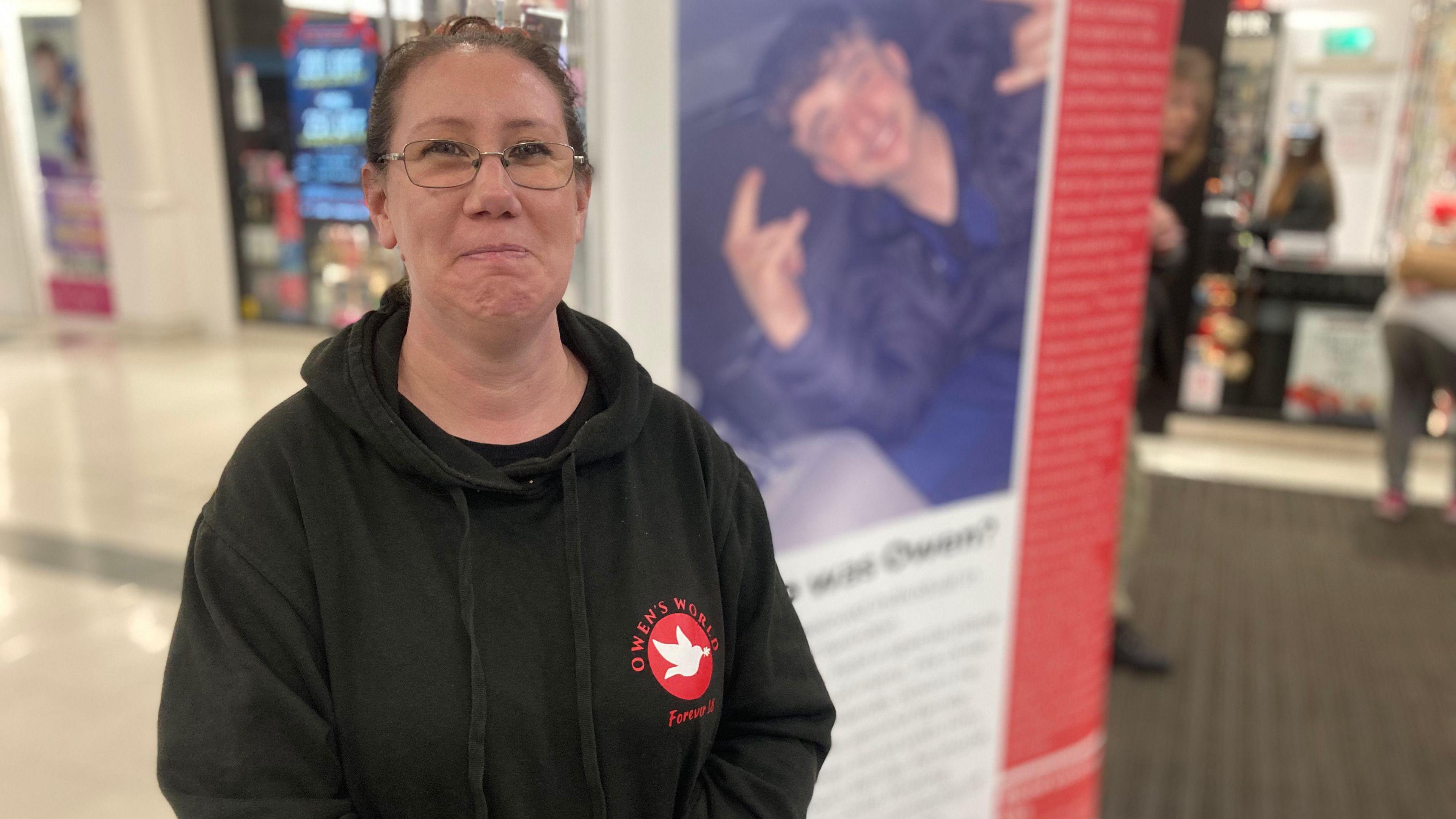 Joanne Davis. She is pictured in front of a large poster with Owen Dunn's photo on it, although it is in the background and out of focus. Joanne has dark hair which is tied up, glasses, and is wearing a black hoodie with a red logo reading "Owen's World" on the side of the chest. She is looking directly at the camera and smiling. 