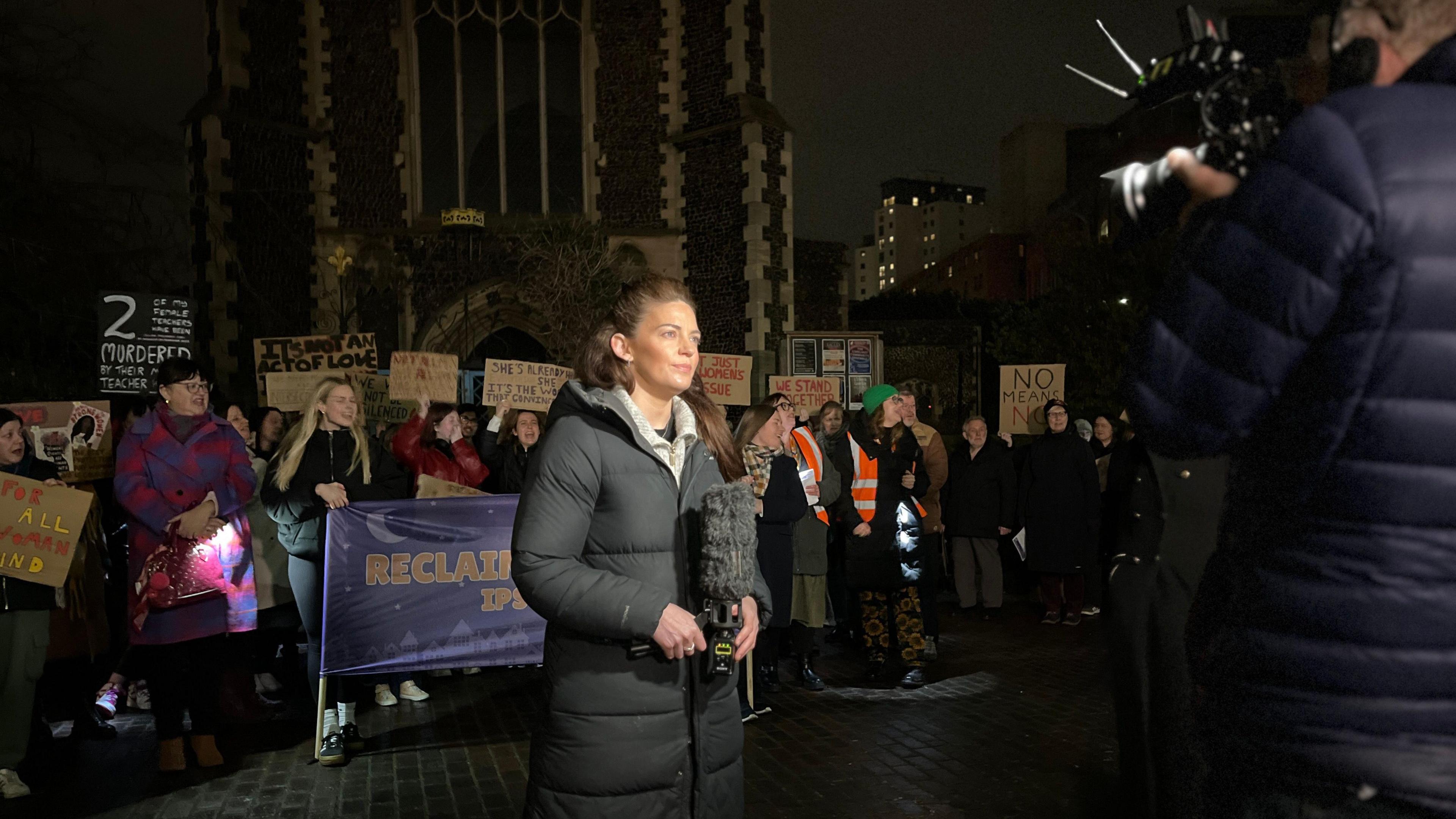 A woman stands in front of a crowd holding a microphone. She is looking at a tv camera.