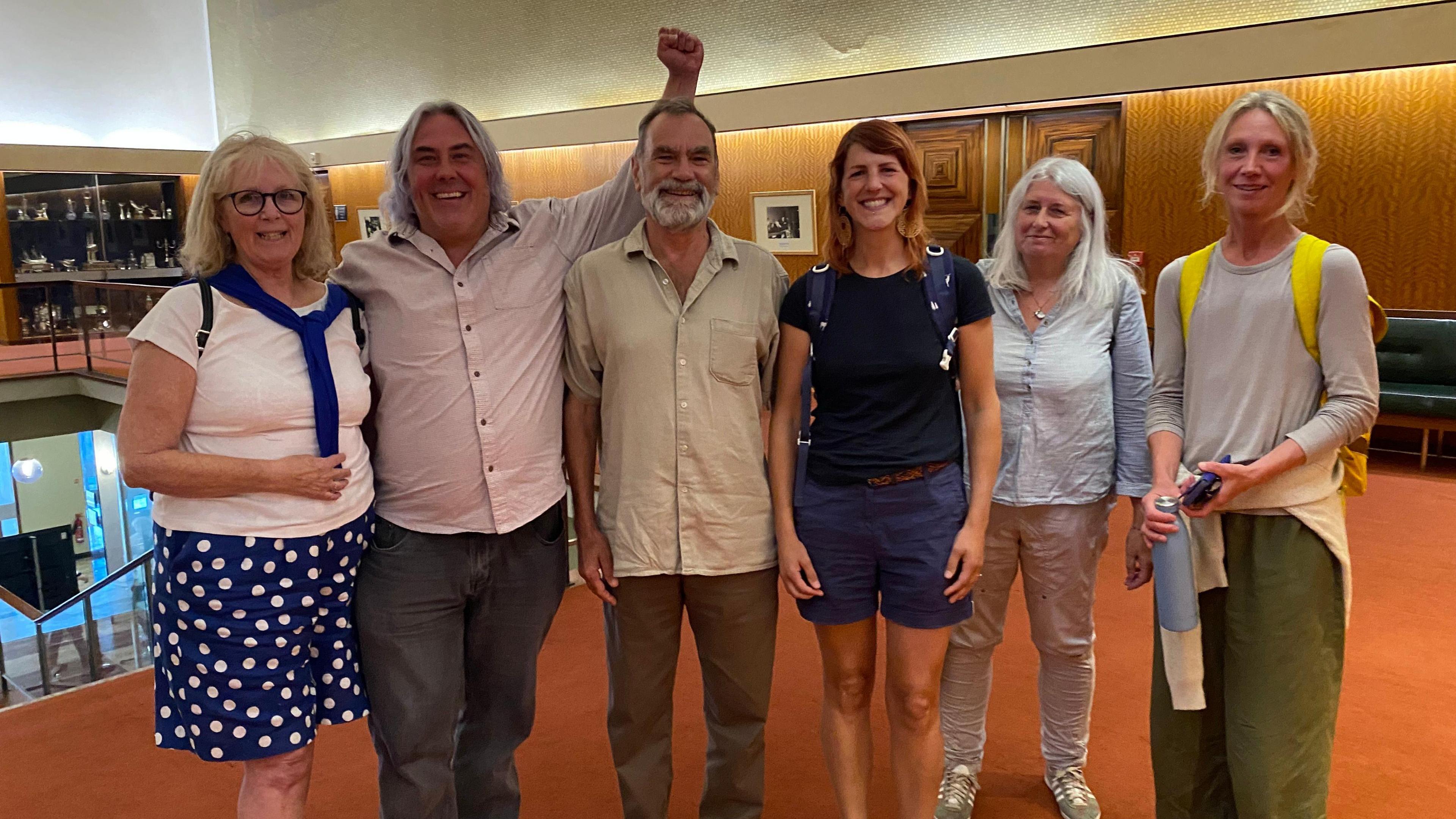 Six campaigners smile and celebrate. They are stood inside Plymouth's Civic Centre building. The carpet is orange. One of the campaigners is holding his left arm in the air.