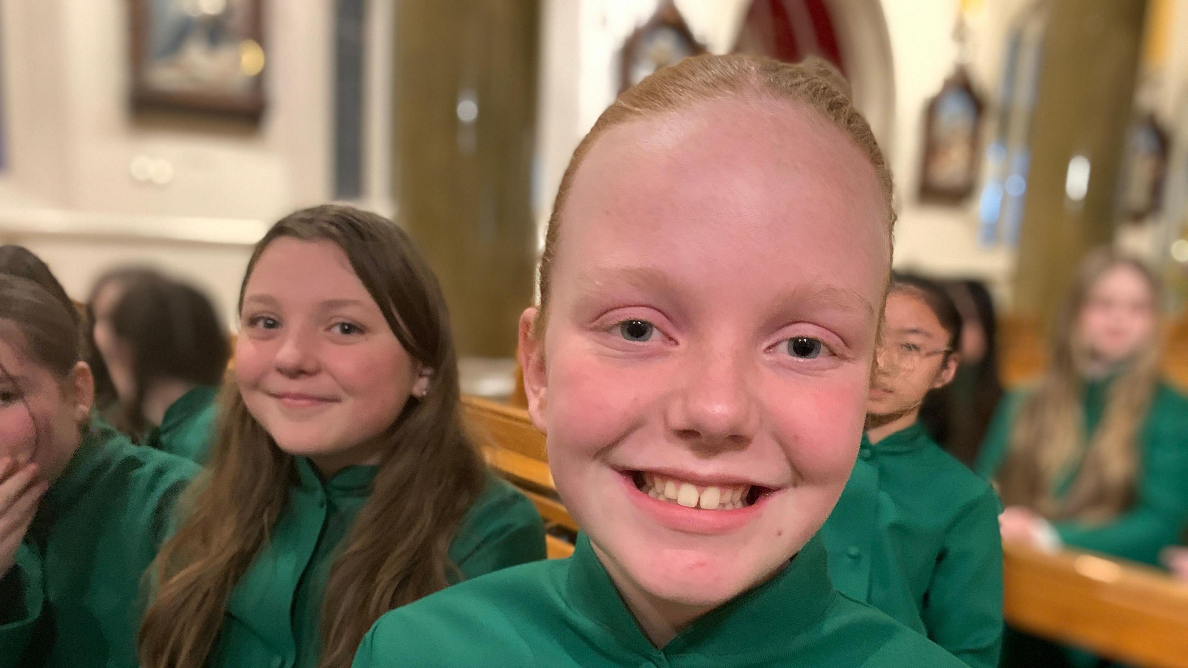 Abbie looking at the camera. She has short auburn hair pulled back behind her head and is wearing a green dress. Beside her a girl with long brown hair looks towards the camera.