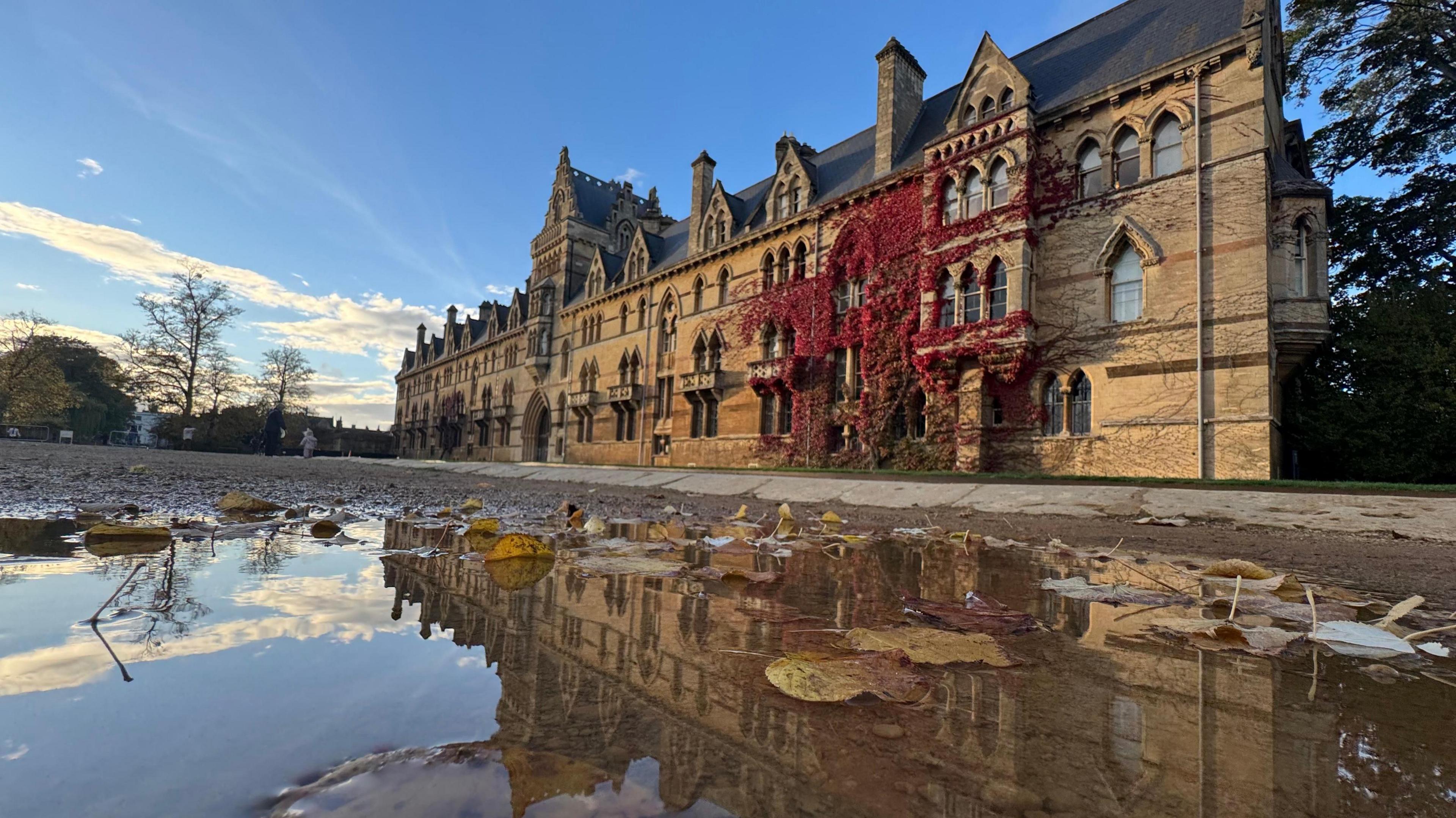 A long, historical building of at least three storeys is reflected in a puddle in low sunshine. The brown walls and arched windows are partly covered by a red climbing plant.